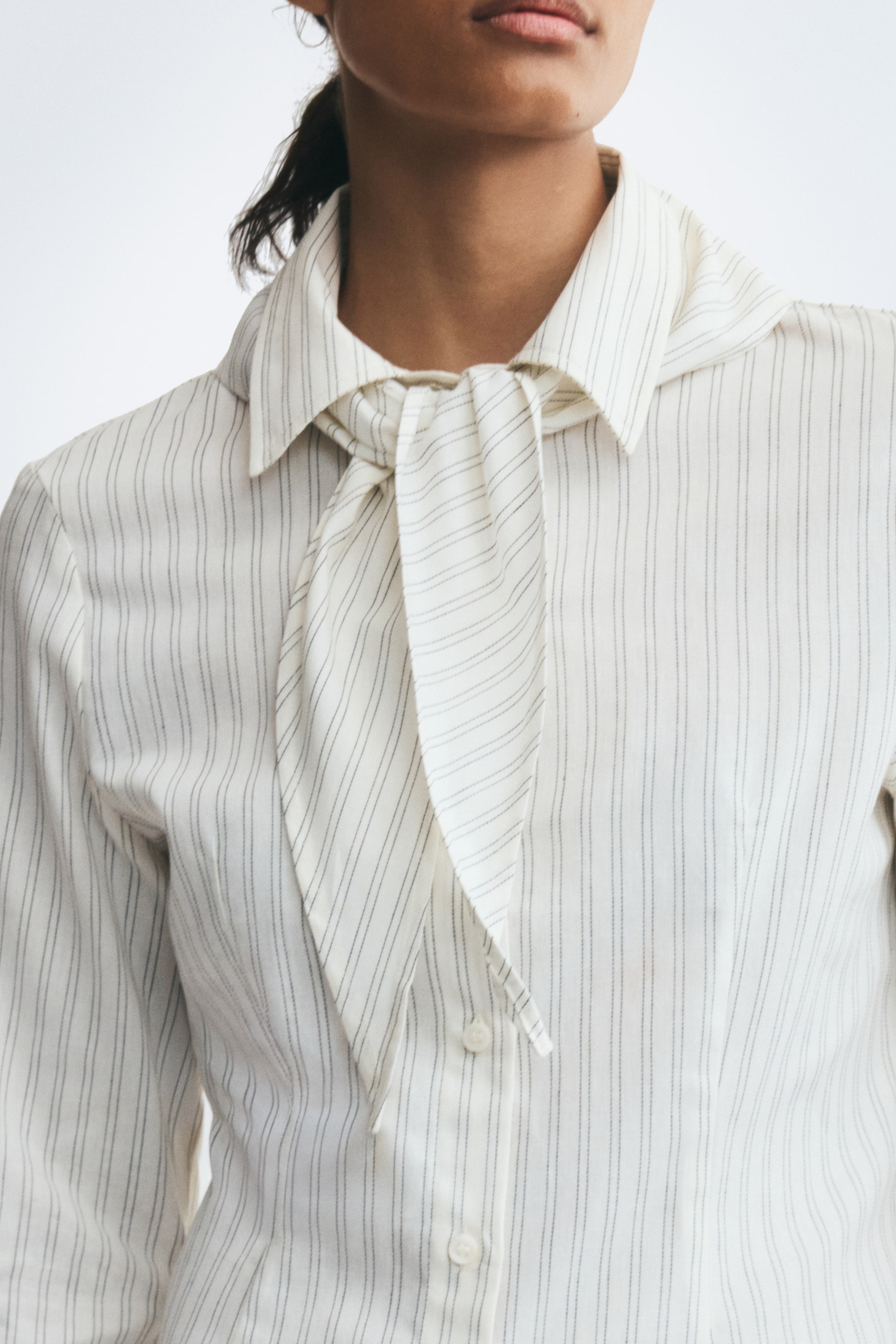 View larger image: The model is wearing a striped, off-white cotton-weave shirt with a pointed collar, a matching scarf tied at the neck, and buttons down the front.