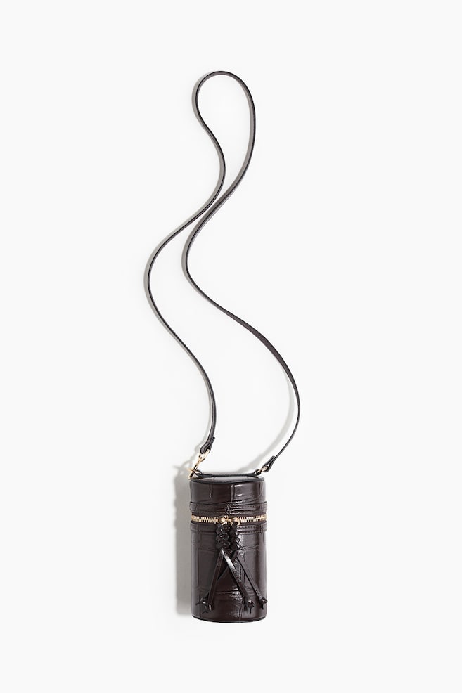 A dark brown, cylindrical bag with a crocodile-embossed texture is facing front, featuring a gold-tone zipper running horizontally around its mid-section with braided leather tassel pulls. A thin, dark brown shoulder strap is attached to the top with gold-tone hardware. - 1