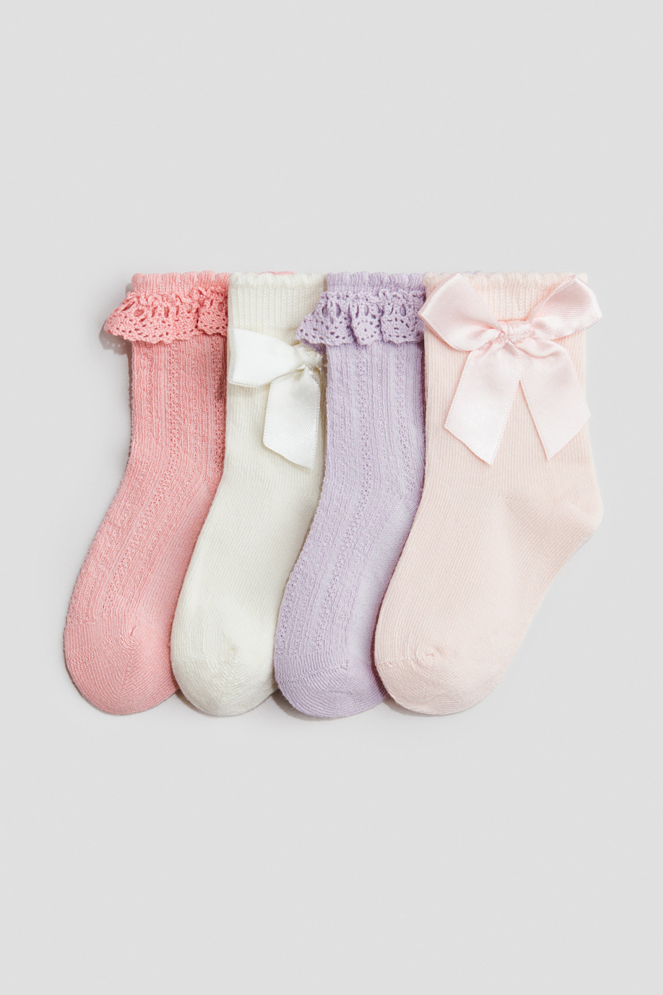 Ampliar la imagen: Cuatro calcetines de tejido fino están tendidos, mostrando su vista frontal. Dos calcetines, uno rosa y otro lavanda, presentan delicados puños con volantes de encaje y diseños de tejido calado en sus piernas, mientras que los otros dos, uno crema y otro rosa claro, ostentan lazos de satén atados en sus puños frontales.