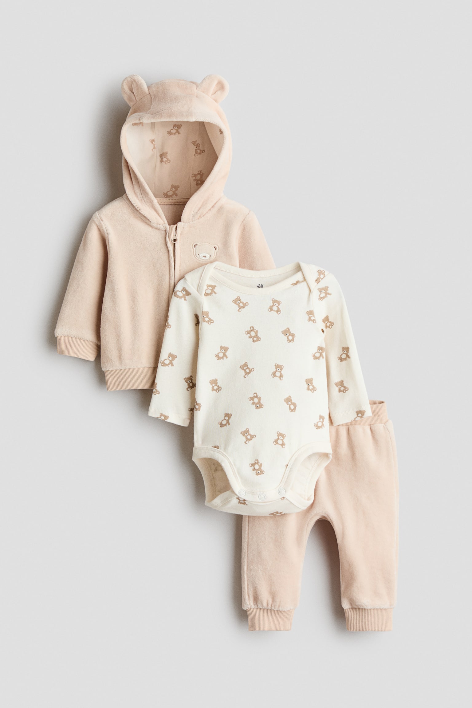 A three-piece baby clothing set features a pale peach zip-through hoodie, with bear ears on the jersey-lined hood and bear print lining, a front zip fastening, an embroidered bear face, and ribbed cuffs and hem. A cream long-sleeved bodysuit, with an all-over small bear print, lapped shoulders, and visible press studs, is paired with matching pale peach joggers, which feature a ribbed waist and hems. - 1