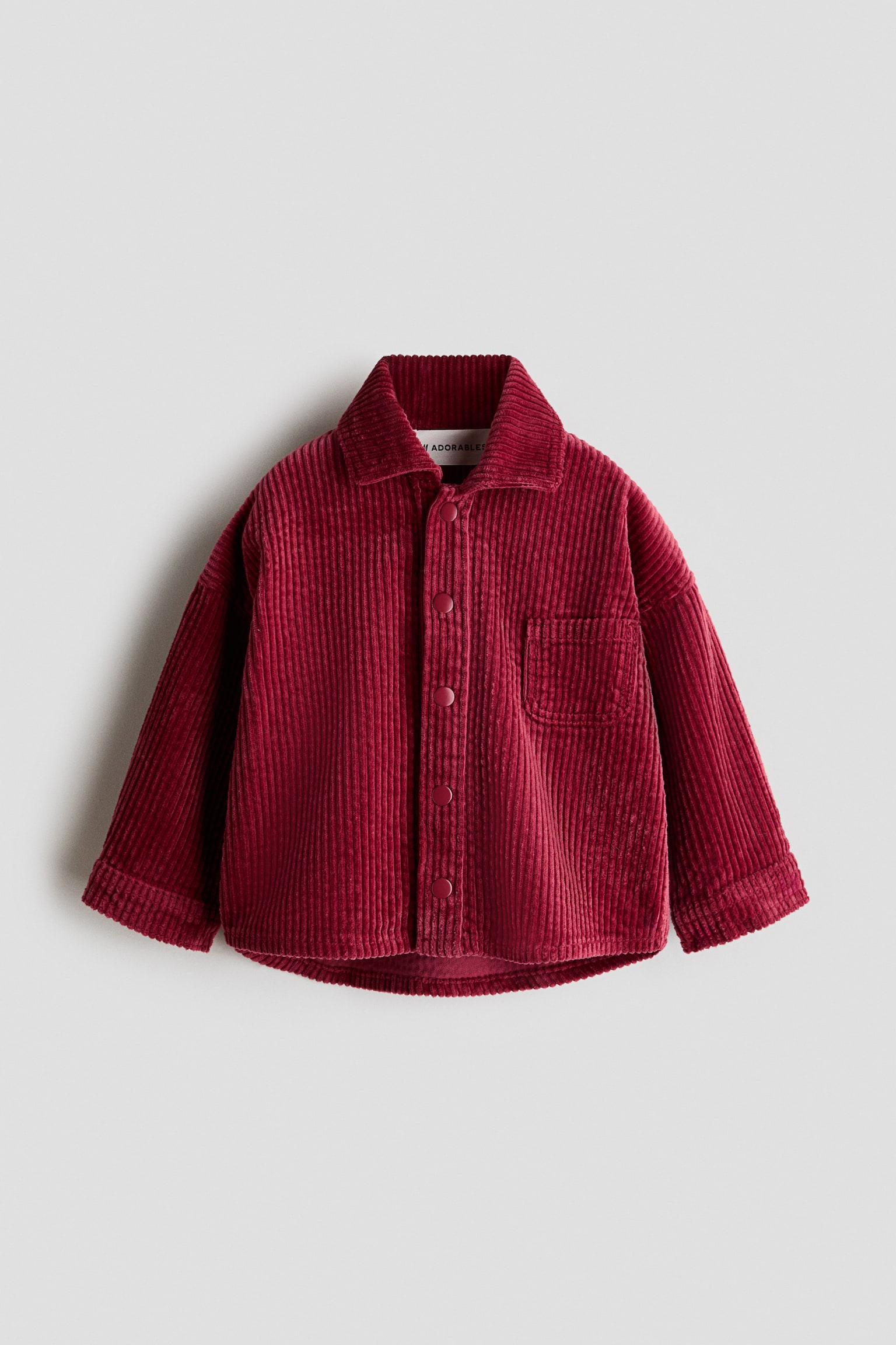 A burgundy corduroy overshirt is shown from the front, featuring a classic point collar, a front snap-button fastening with colour-matched buttons, and a single chest patch pocket. The shirt features dropped shoulders, long sleeves with folded cuffs, and a curved hem.
