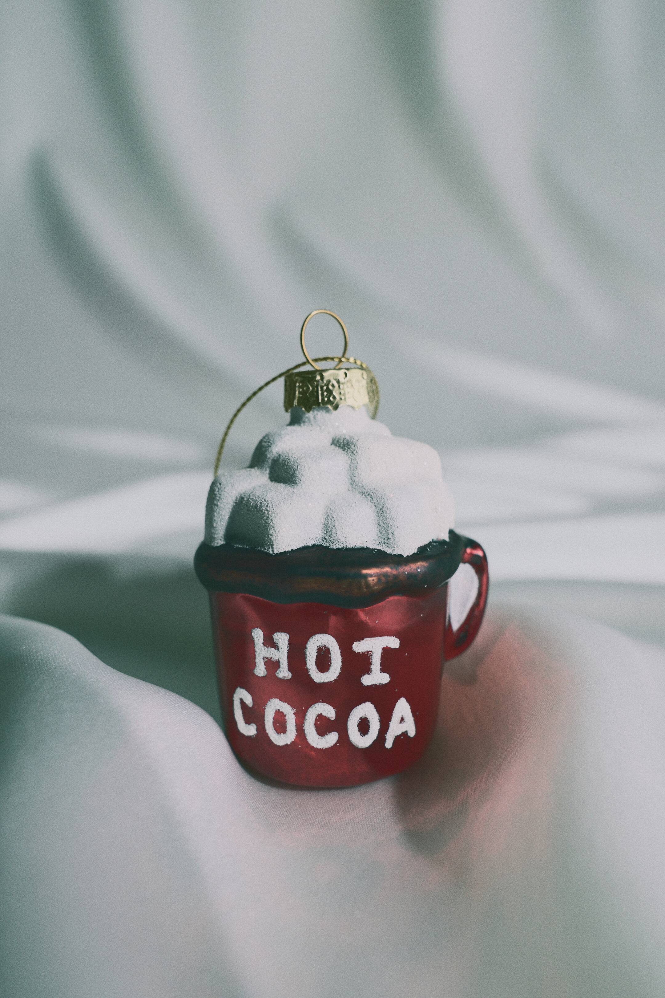 Büyük resmi görüntüle: Kırmızı bir kupa şeklinde cam bir yılbaşı süsü öne dönük duruyor ve üzerinde beyaz blok harflerle "HOT COCOA" yazısı yer alıyor. Süs, dokulu, beyaz çırpılmış krema tarzı bir üst süslemeyi sergiliyor ve tepesinde asma halkalı altın renkli bir kapak bulunuyor.