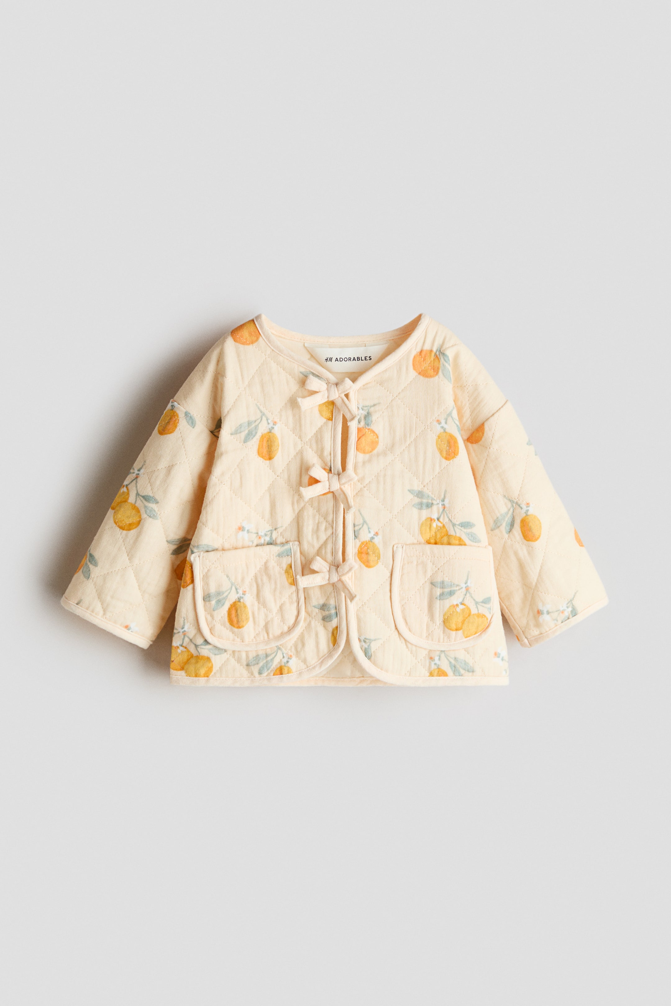 Ver imagem maior: Um casaco acolchoado amarelo-claro voltado para a frente, com costura em losango, exibe um padrão de frutas cítricas laranja e folhas verdes. É desenhado com uma gola redonda, três fechos de laço de cor clara na frente e dois bolsos arredondados aplicados.