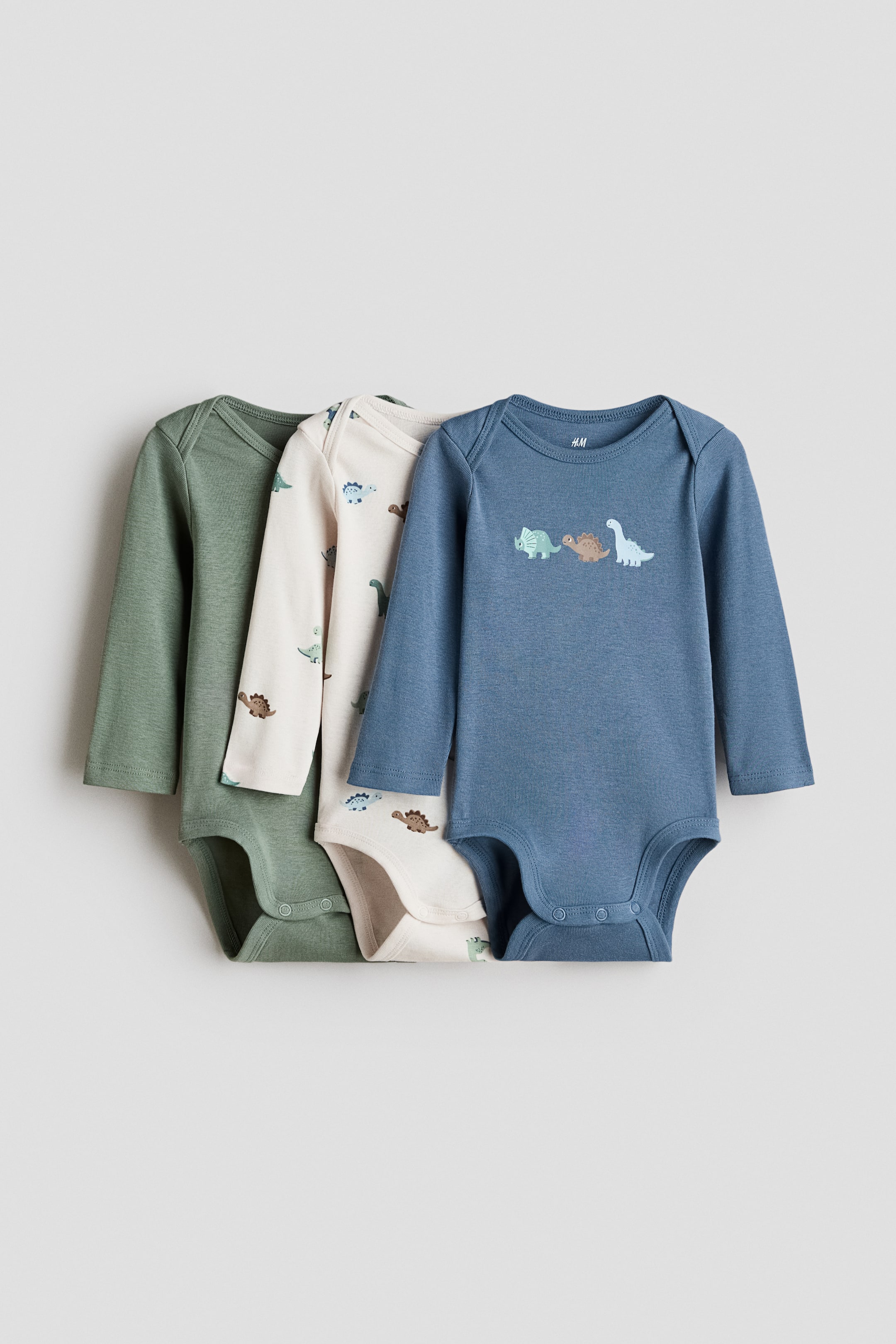 View larger image: Three long-sleeved baby bodysuits are arranged in layers from front to back, all facing forward. The front blue bodysuit showcases three small dinosaur graphics on its chest, while the middle cream bodysuit boasts an all-over dinosaur print. The back green bodysuit features a solid color. All three bodysuits come with round necklines and snap closures at the crotch.
