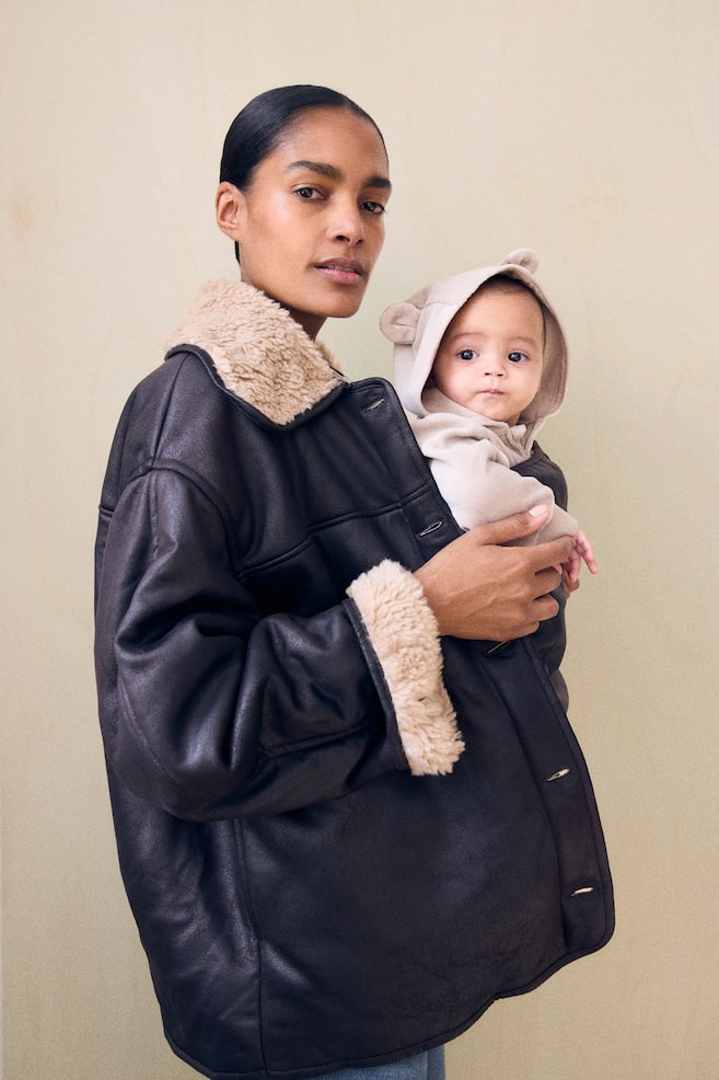 A dark brown shearling jacket is being worn by the model, featuring a plush beige shearling collar and cuffs. The jacket is being held partially open to envelope a baby wearing a hooded beige romper who is being carried against the model's chest. - 1