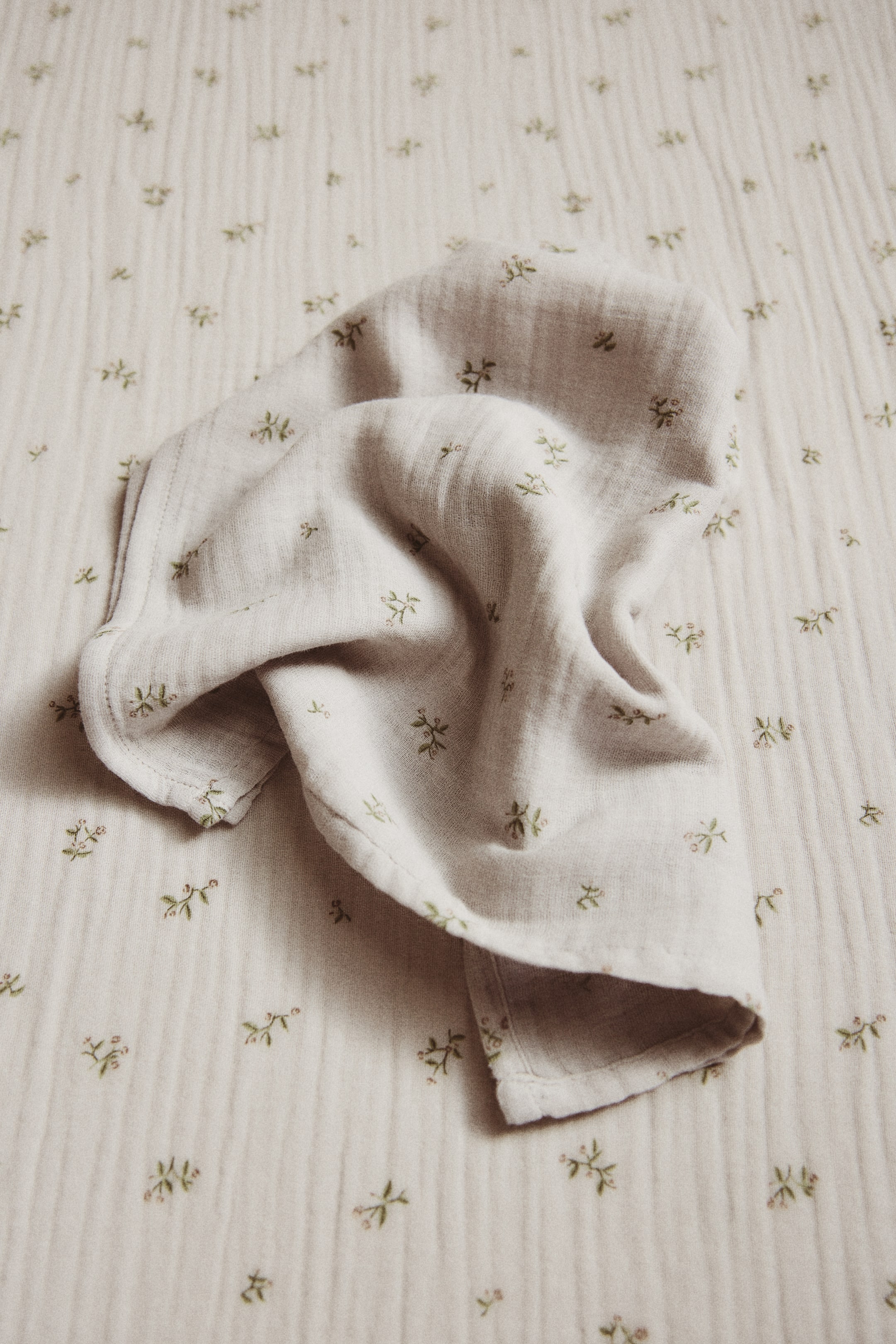 Agrandir l'image: Un tissu en mousseline de coton beige clair est partiellement plié et froissé, affichant un motif dispersé de petites fleurs vertes et roses.