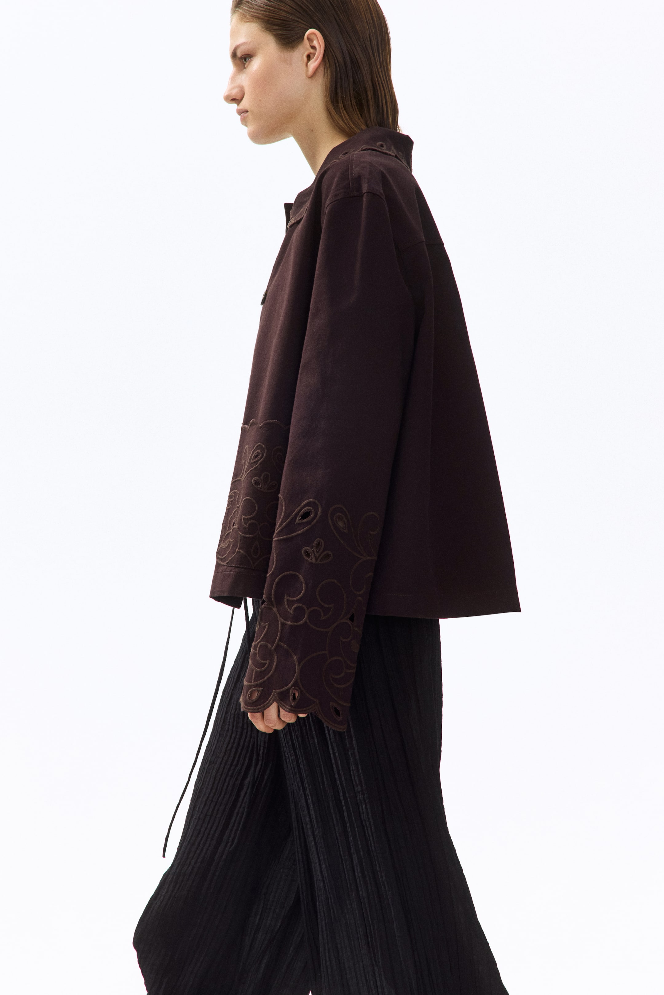 View larger image: Presenting a dark brown jacket from a side profile, it showcases wide, long sleeves adorned with intricate swirling cut-out embroidery and matching stitching at the cuffs. The jacket is styled with a relaxed, cropped, and boxy fit.