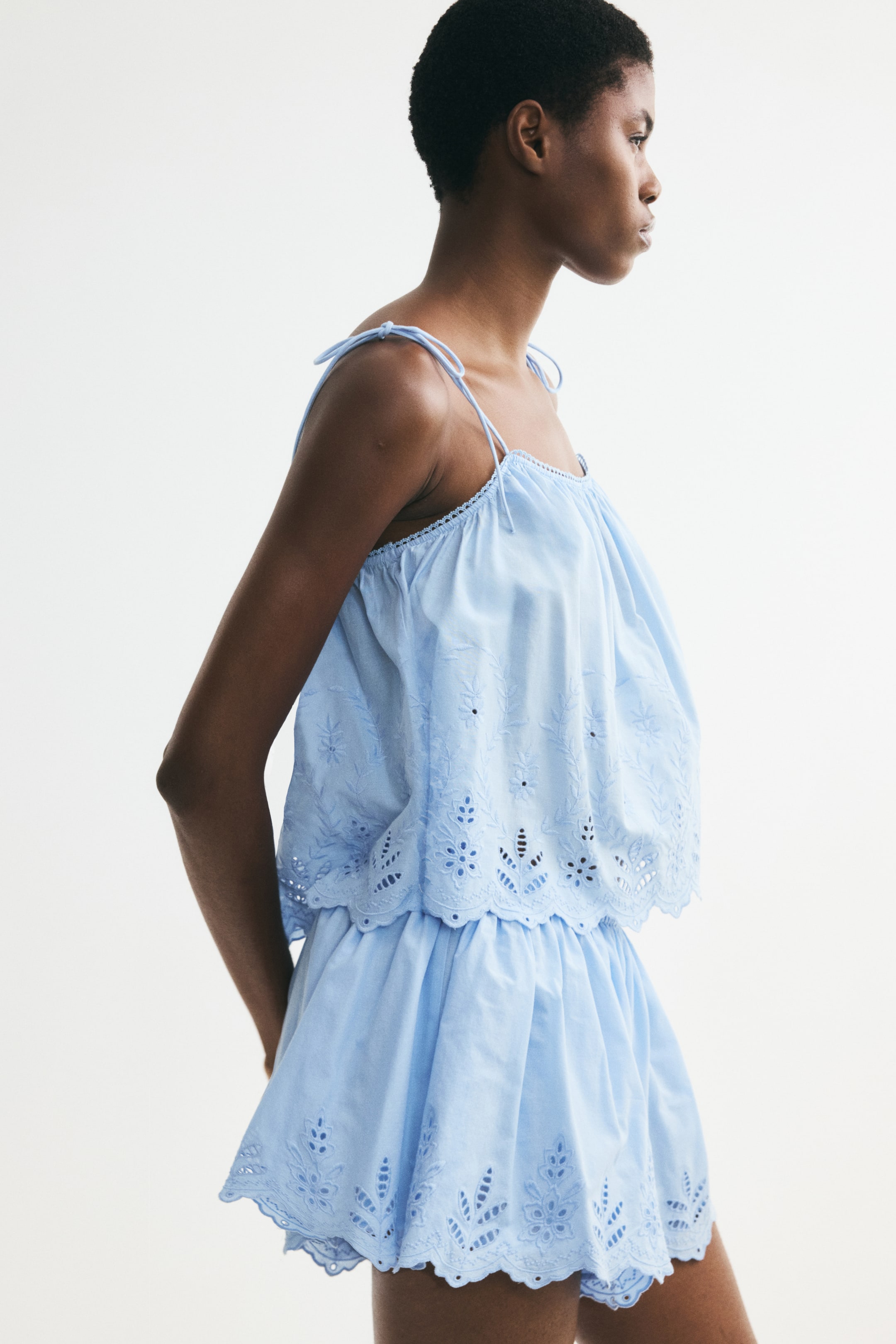 View larger image: A light blue two-piece set is showcasing its side profile, featuring a camisole top with thin tie straps, a gathered neckline, and a loose, tiered body with broderie anglaise and a scalloped hem. The matching shorts complement the top with a gathered waist, broderie anglaise, and a scalloped hem.