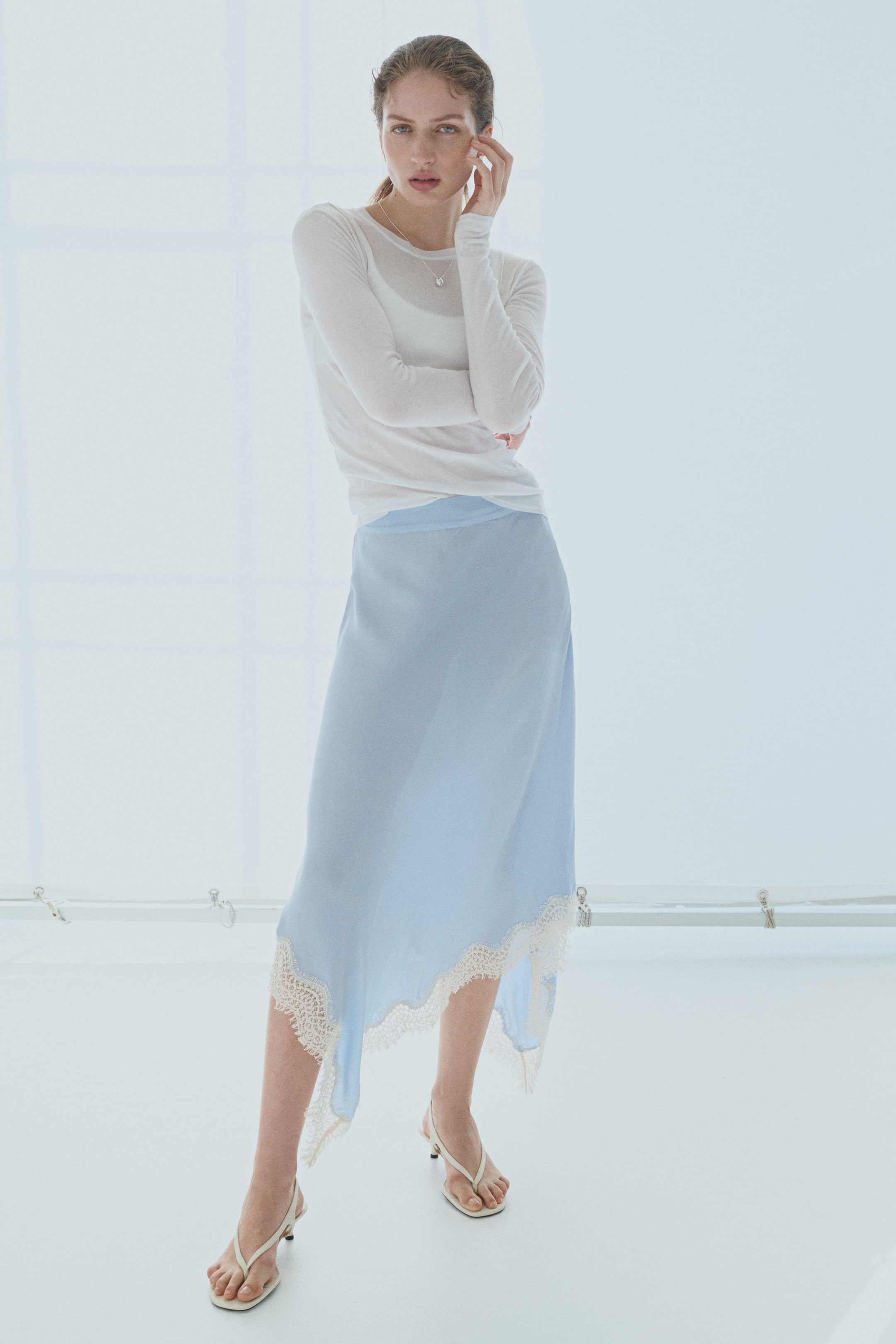 View larger image: A light blue midi skirt is flowing from a slight front-left angle, featuring an asymmetrical hemline adorned with cream lace trim. The lace trim is showcasing a scalloped edge and intricate patterns.