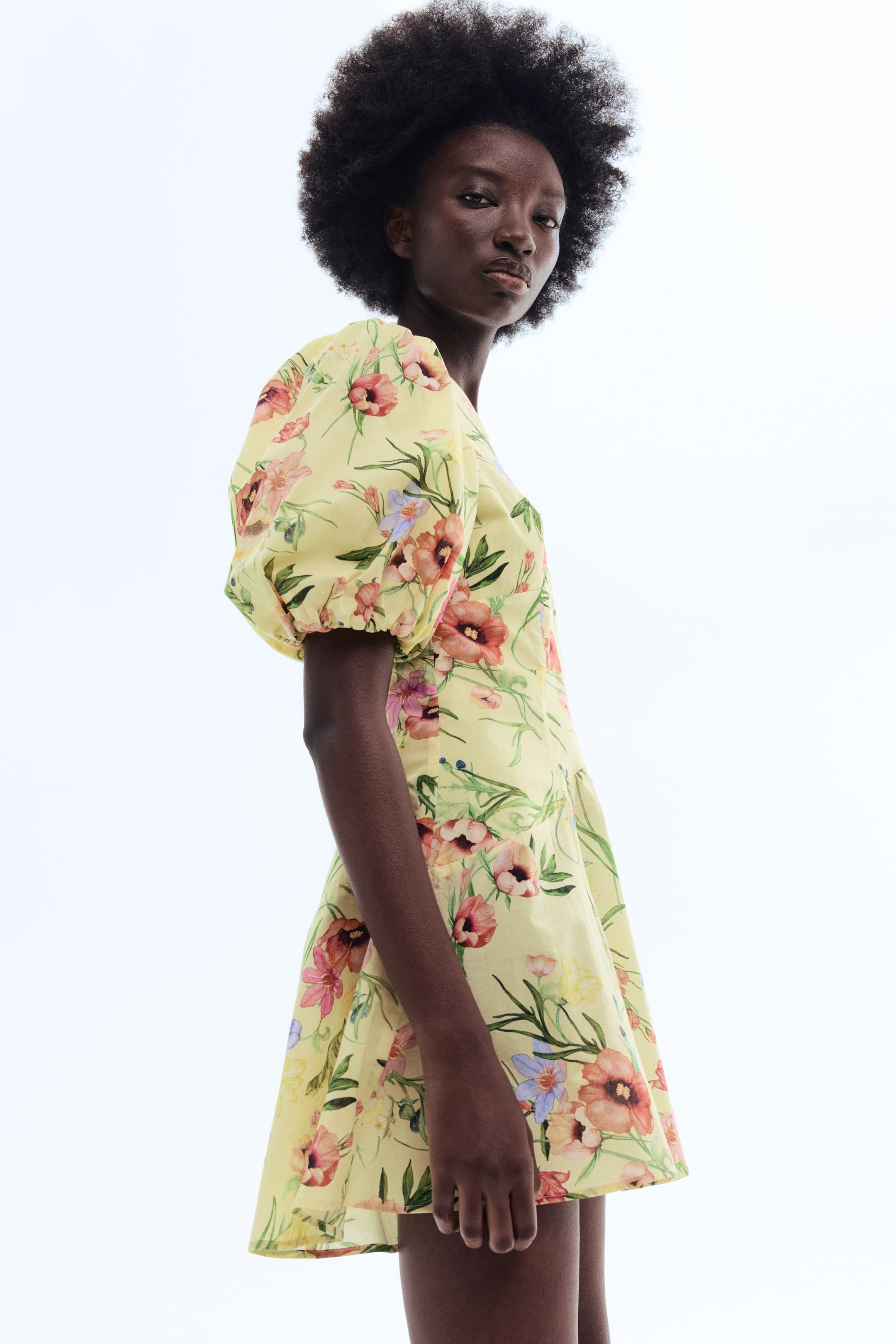 View larger image: A light yellow floral dress featuring puffed short sleeves with gathered cuffs and a square neckline. It showcases a pattern of pink, red, orange, and blue flowers with green leaves, presenting an A-line skirt from a side-front angle.