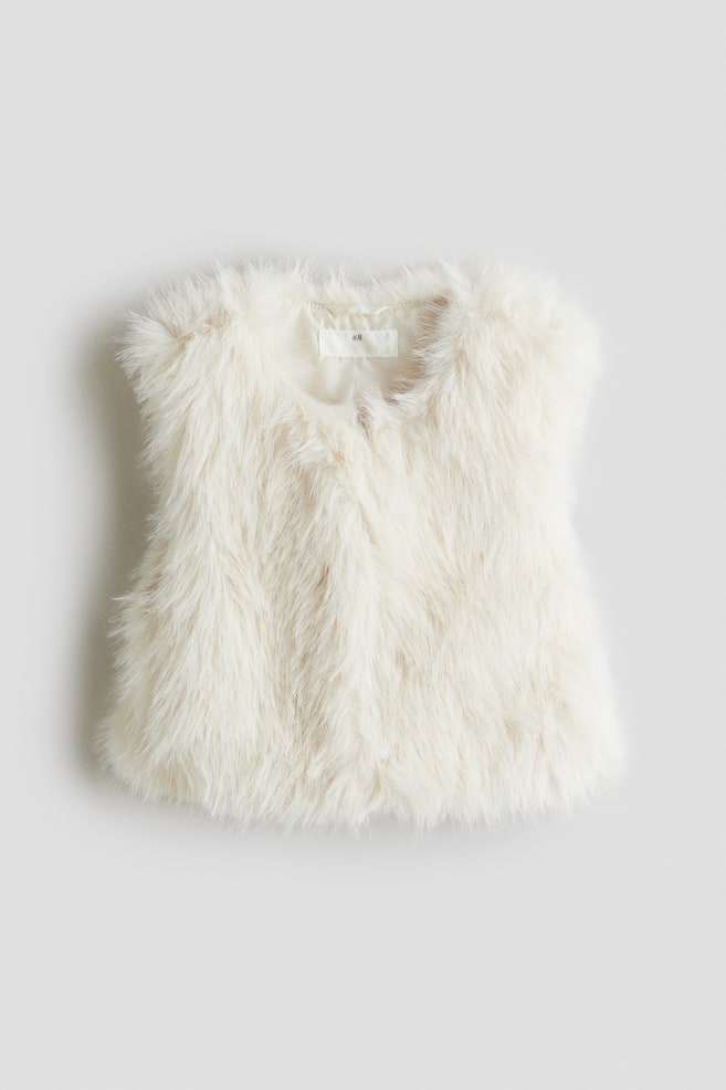 Un gilet en fausse fourrure de couleur crème est présenté de face, avec une texture longue et hirsute et un col rond sans col. Une petite étiquette blanche est visible sur la doublure intérieure à l'arrière du cou. - 1