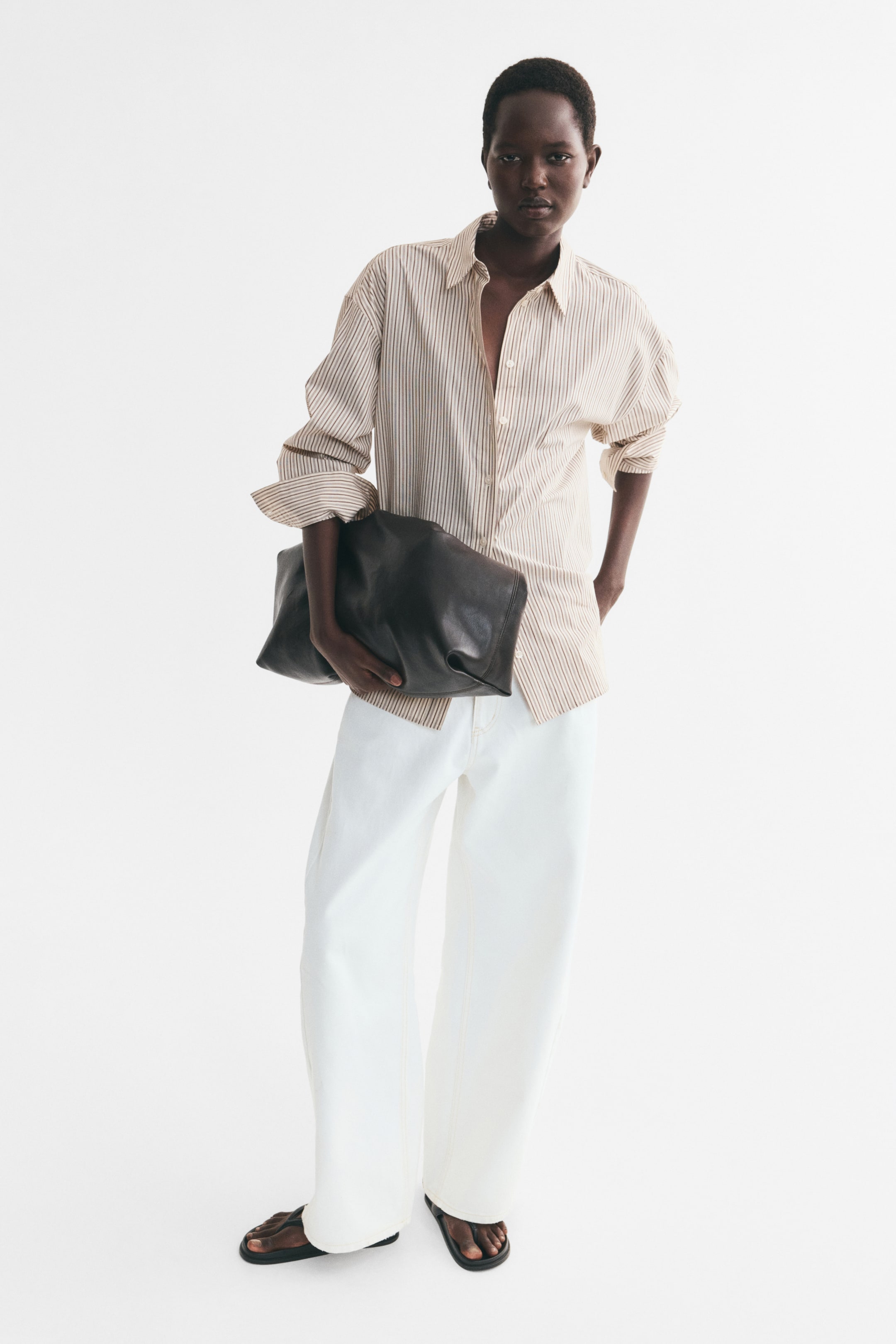 View larger image: An oversized beige-and-white vertically striped shirt is worn by the model, styled with white wide-leg trousers. The shirt features a collar, dropped shoulders, and a button-front closure, with the sleeves rolled up. The model is holding a large dark leather clutch bag under one arm.