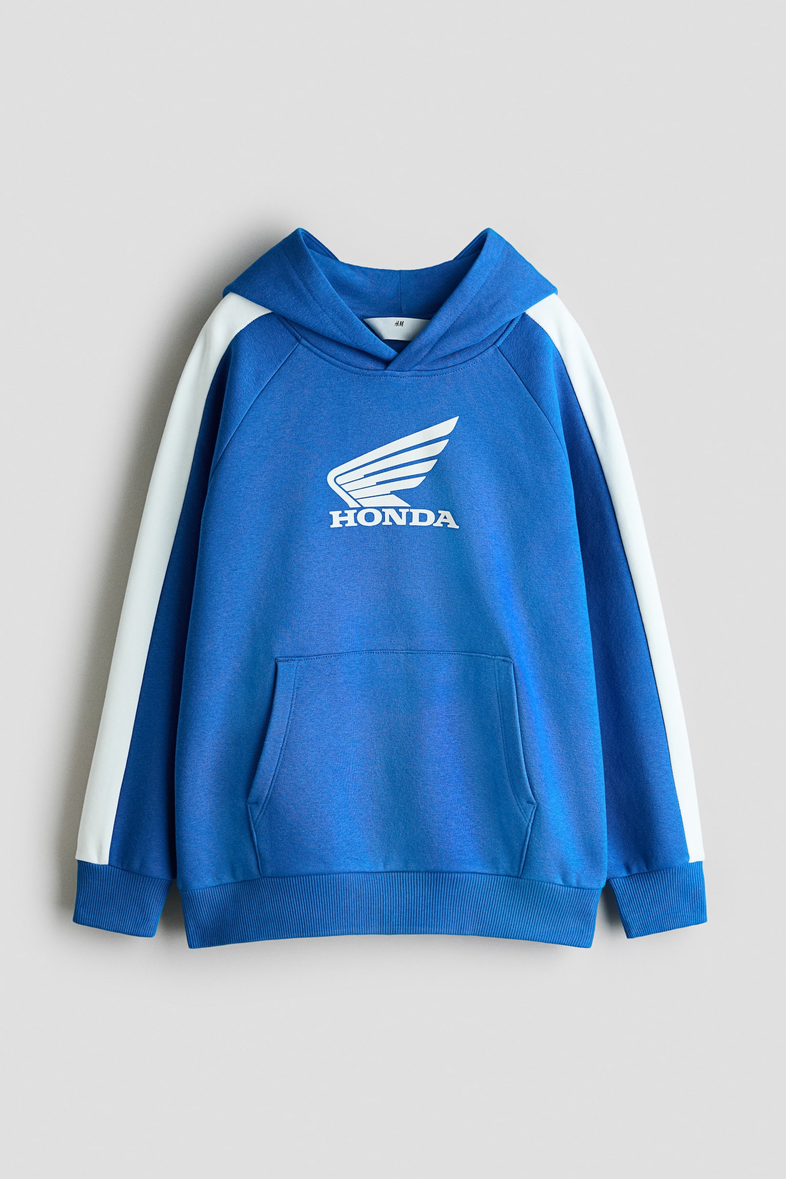 A blue fleece hoodie is shown in a front view, featuring a white Honda wing logo printed on the centre chest. The design highlights a wrapover hood, long raglan sleeves with contrasting white stripes down the sides, a spacious kangaroo pocket, and ribbed detailing at the cuffs and hem.