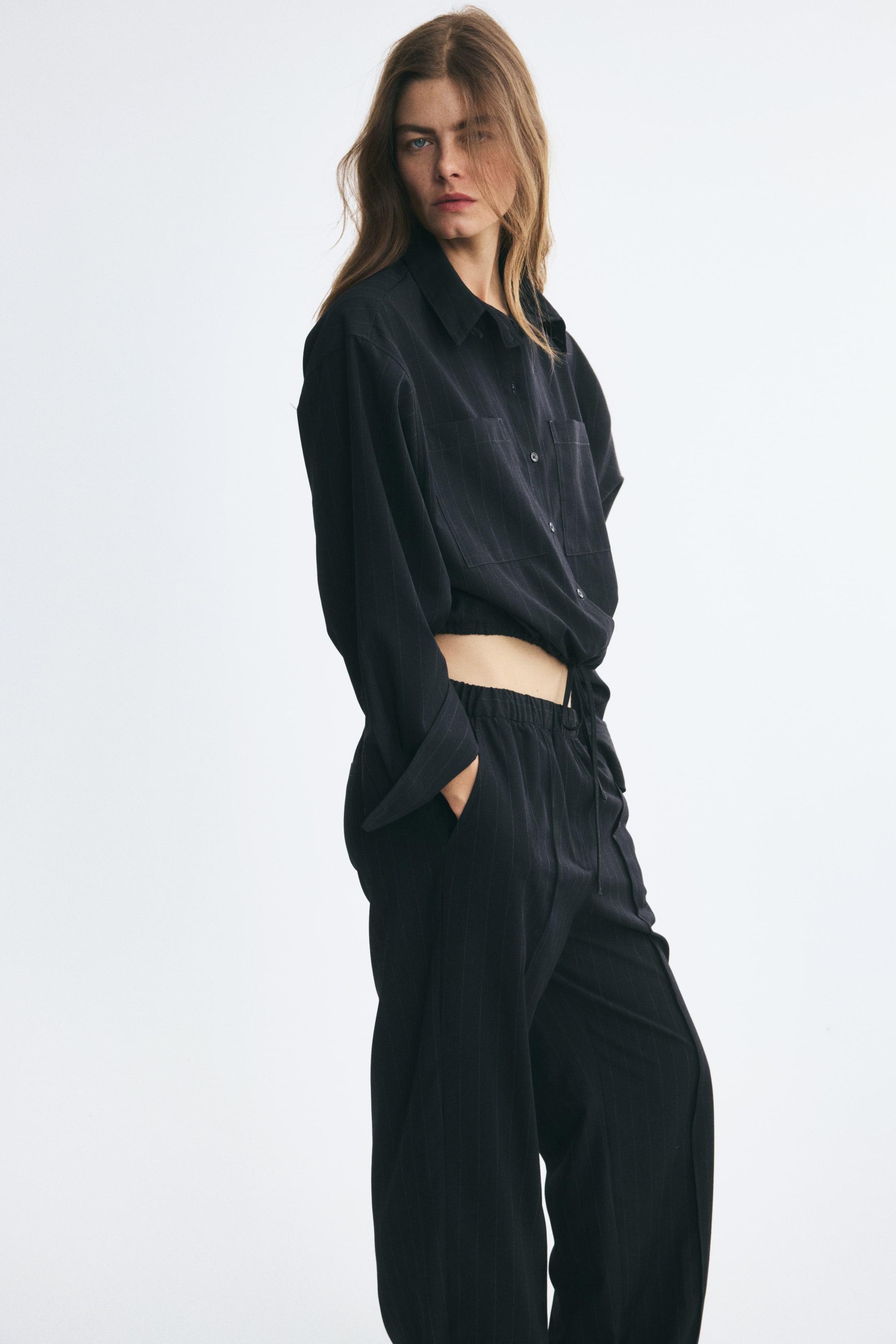 Ampliar la imagen: La modelo lleva una camisa negra de rayas finas de corte oversize, combinada con pantalones a juego. La camisa presenta cierre frontal de botones, bolsillos de parche en el pecho, hombros caídos y un dobladillo con cordón ajustable ceñido a la cintura.