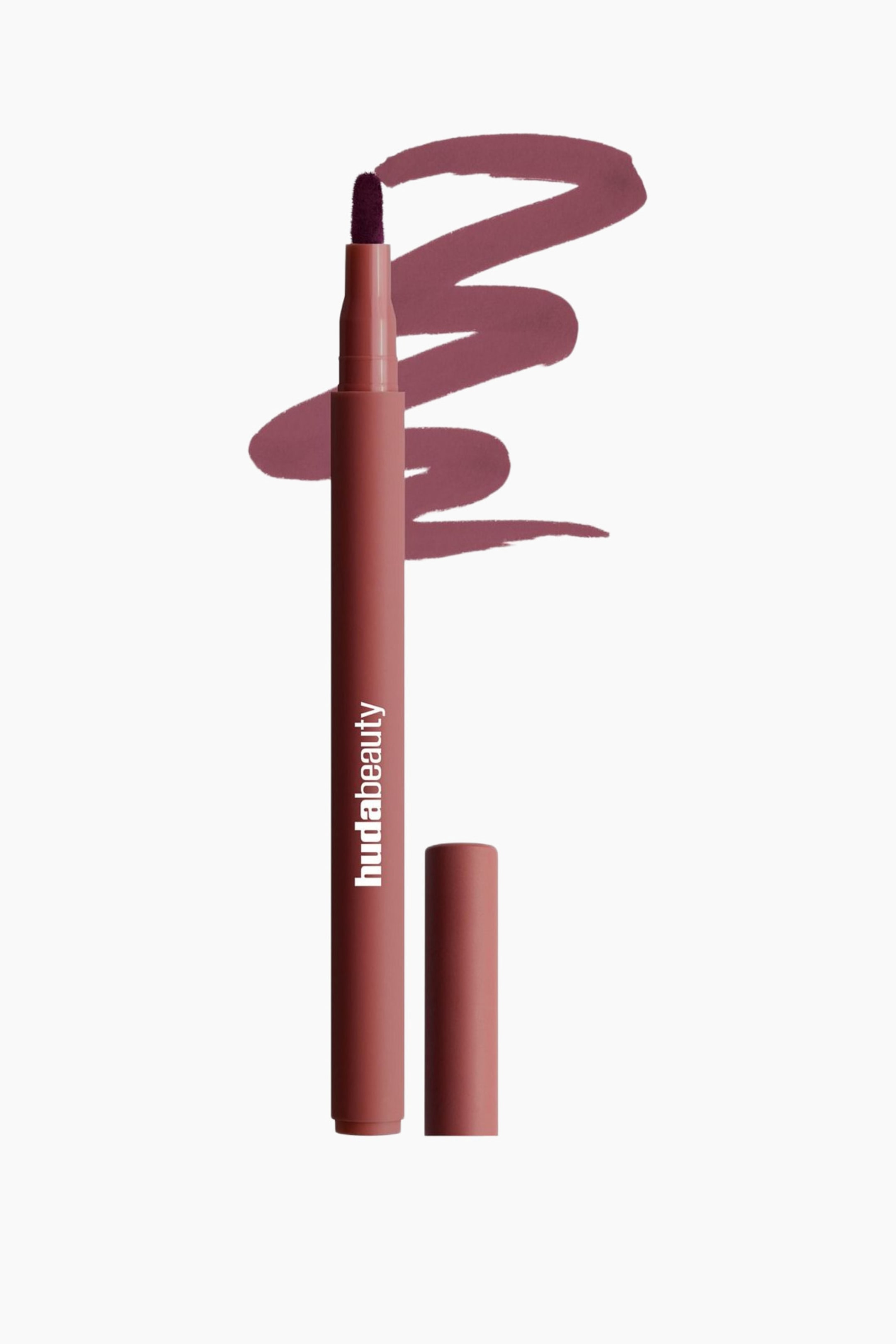 Visa större bild: En Huda Beauty-läppmarkerare står upprätt och har en rödbrun hylsa med "hudabeauty" tryckt på den samt en mörklila filtspets. Dess matchande kork är placerad bredvid markörens bas, och ett rödbrunt färgprov böjer sig bakom produkten.