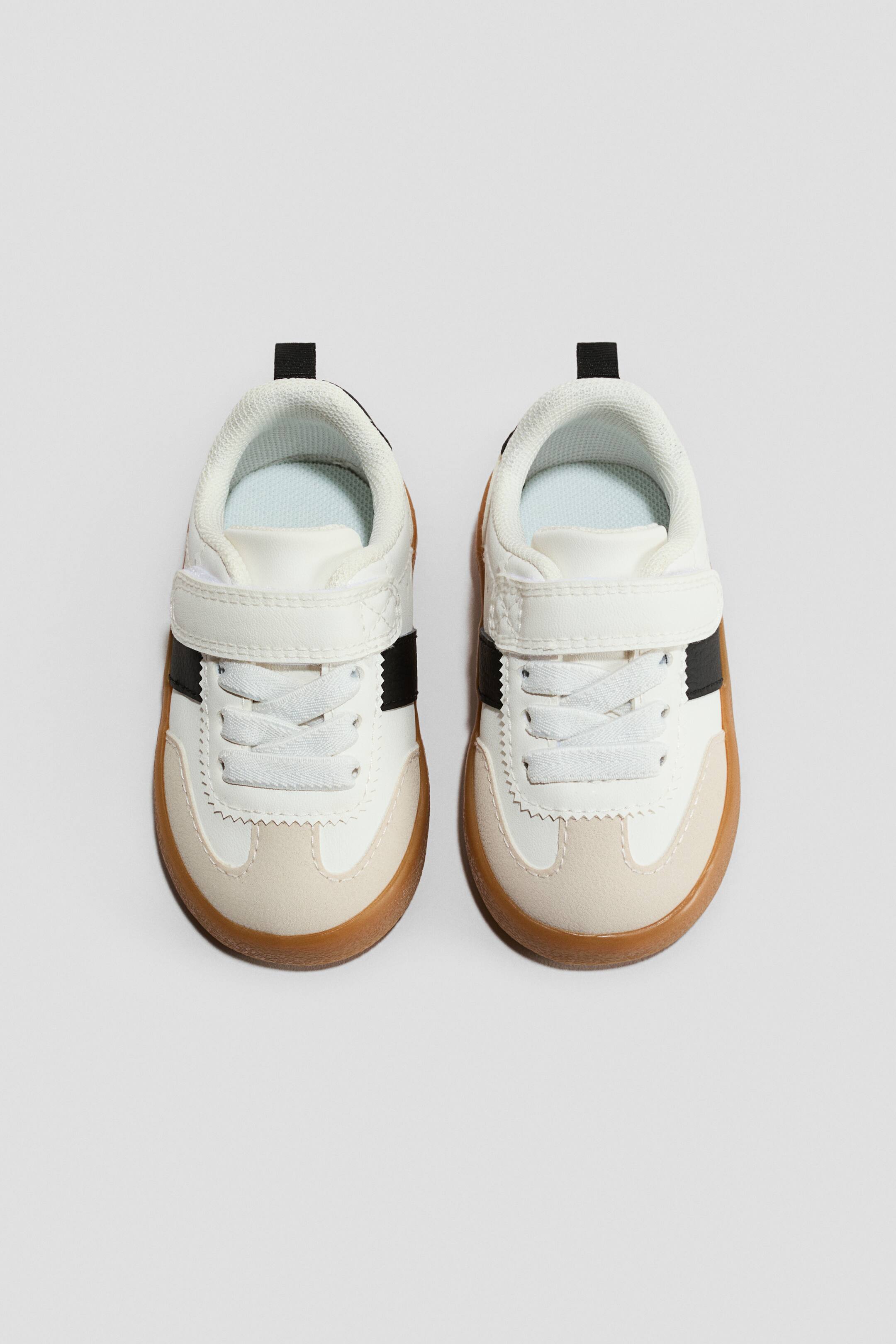 Ampliar la imagen: Los entrenadores están de frente, con partes superiores blancas con punteras de tipo gamuza beige y paneles laterales, y cordones blancos visibles debajo de una correa de gancho y lazo blanca. Una raya negra acentúa el lado, una lengüeta de tracción negra está presente en el talón, y una suela de color goma subraya la parte inferior.