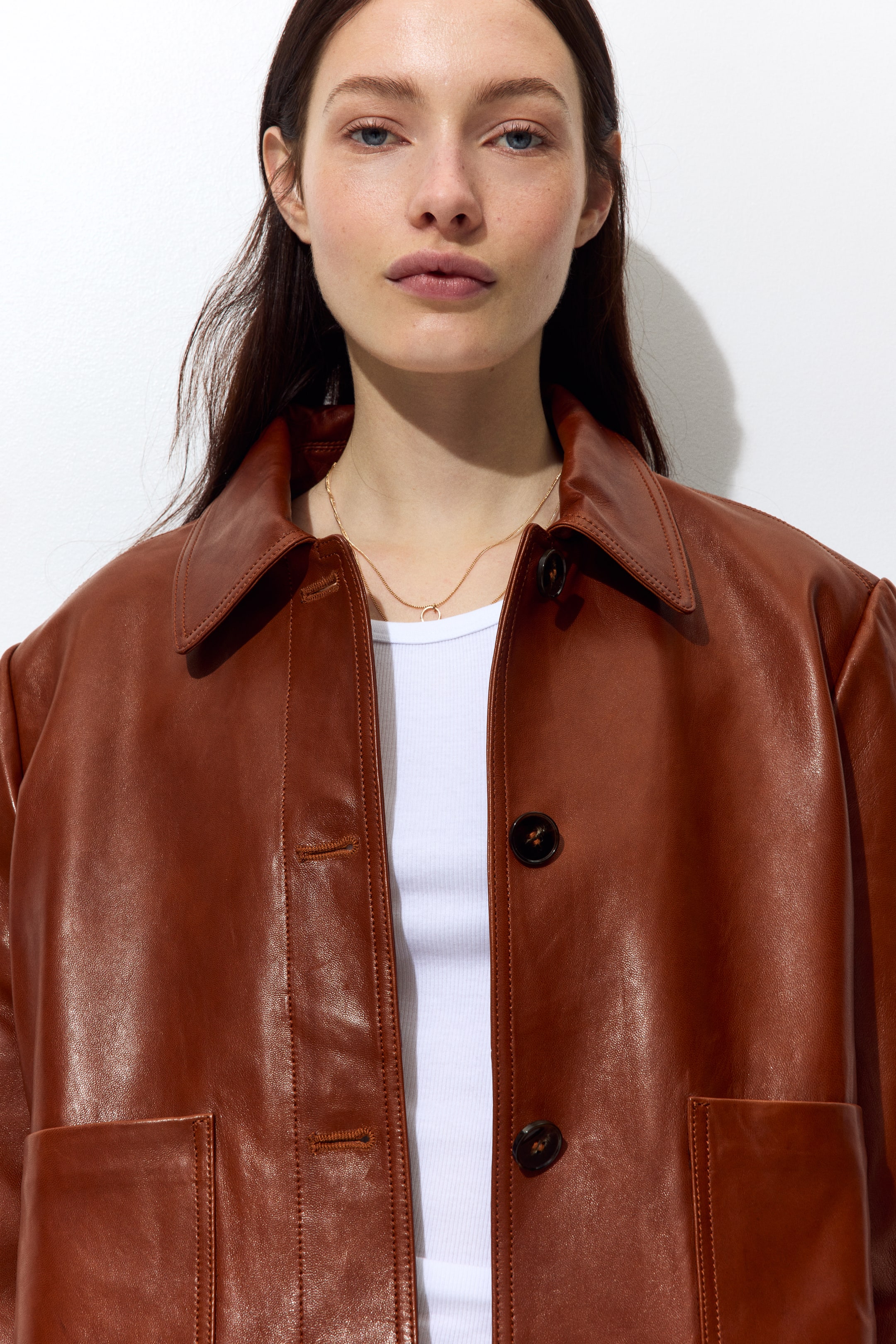 View larger image: A brown leather jacket is showcased, featuring a classic pointed collar, a button-down front with dark brown mottled buttons, and two front patch pockets. It's styled open, revealing a white ribbed top and a thin gold chain with a small circle pendant.