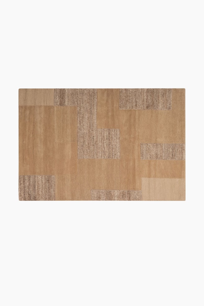 Een rechthoekig tapijt wordt van bovenaf weergegeven, met een geometrisch patchwork ontwerp. Het laat verschillende tinten beige en lichtbruin zien, met delen van gladde, geribbelde en getextureerde poolafwerkingen. - 1