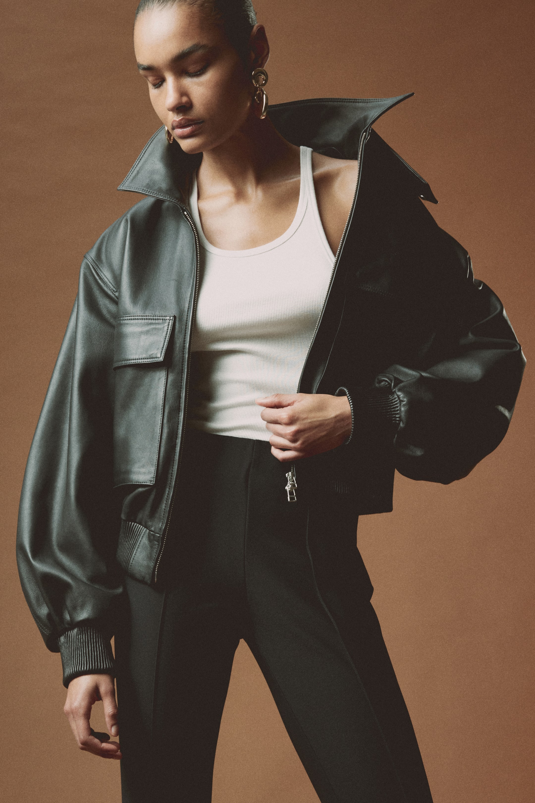 View larger image: The model is wearing black trousers featuring slim legs and stitched-in creases along the front. She is gently holding the visible zip pull at the waistband of the trousers.