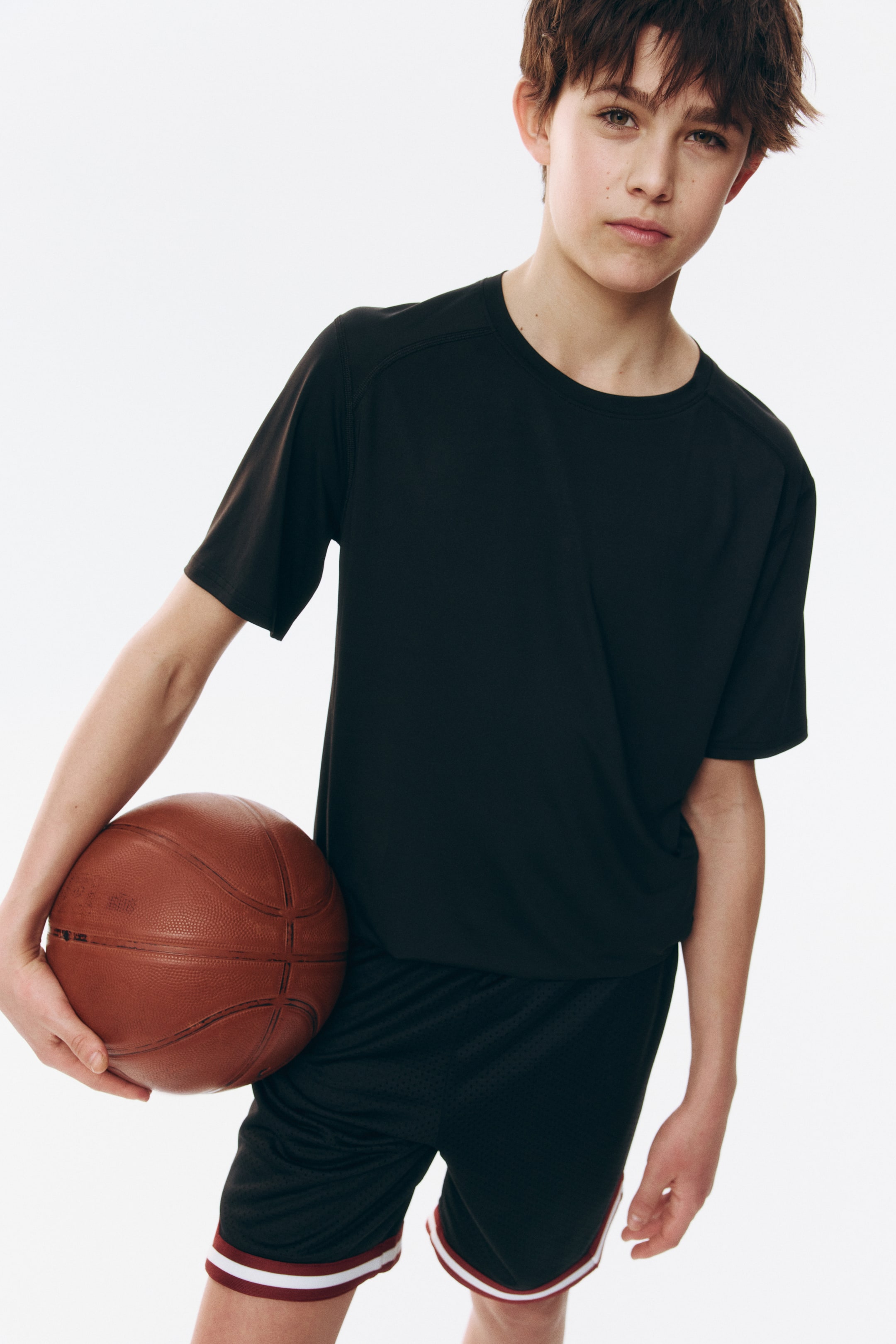 Visa större bild: En svart kortärmad t-shirt med rund hals och raglanärmssömmar bärs. Svarta meshshorts har vit och vinröd randig kant längs benöppningarna.