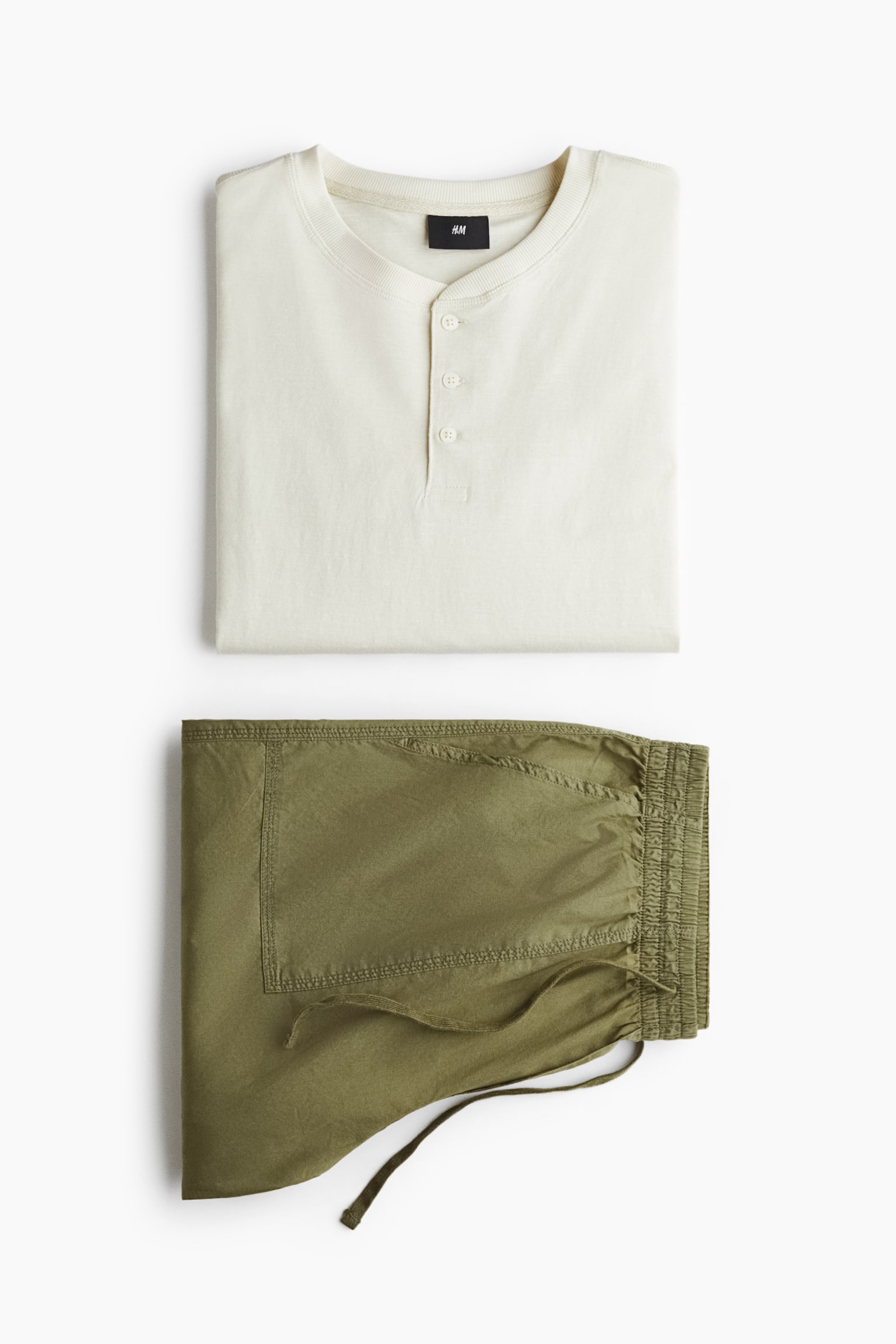 An off-white Henley shirt and olive green trousers are presented folded and facing front as a coordinated set. The shirt features a ribbed round neckline with a three-button placket, while the trousers feature an elasticated drawstring waistband and a stitched side pocket. - 1