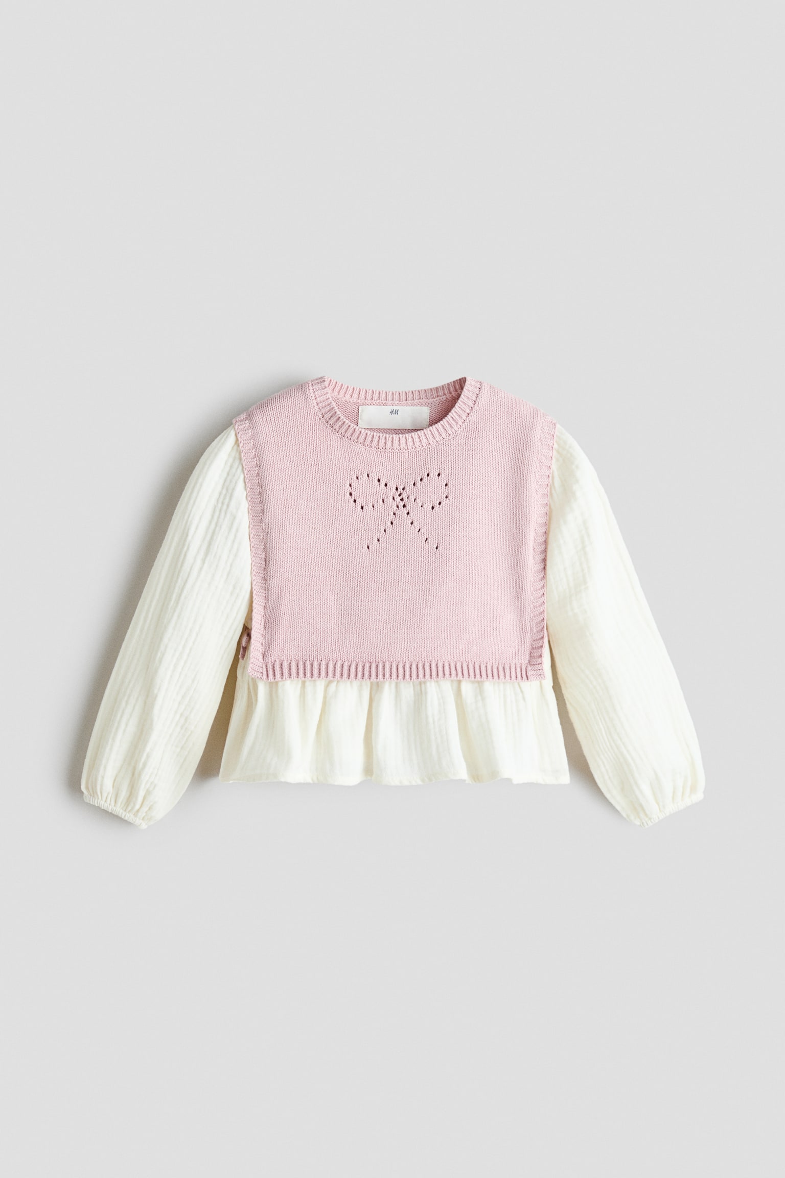 Ein zweiteiliges Kinder-Top ist von vorne zu sehen und besteht aus einer hellrosa Strickweste, die Ã¼ber einer cremeweiÃen Bluse mit Struktur getragen wird. Die rosa Weste zeigt einen gerippten Rundhalsausschnitt und ein Pointelle-Schleifendetail auf der Brust, wÃ¤hrend die darunterliegende Bluse lange Ãrmel mit elastischen BÃ¼ndchen und einen ausgestellten Peplum-Saum aufweist.