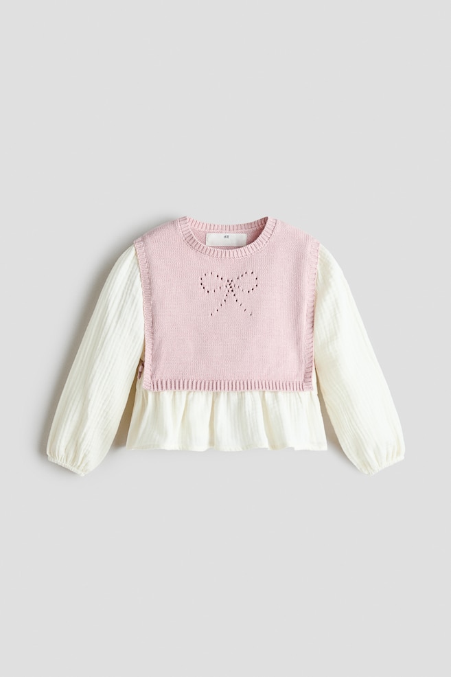 Una maglia due in uno per bambini è mostrata frontalmente, con un gilet in maglia rosa chiaro sovrapposto a una blusa testurizzata bianco sporco. Il gilet rosa presenta un girocollo a coste e un motivo a fiocco pointelle sul petto, mentre la blusa sottostante mostra maniche lunghe con polsini elasticizzati e un orlo peplum svasato. - 1