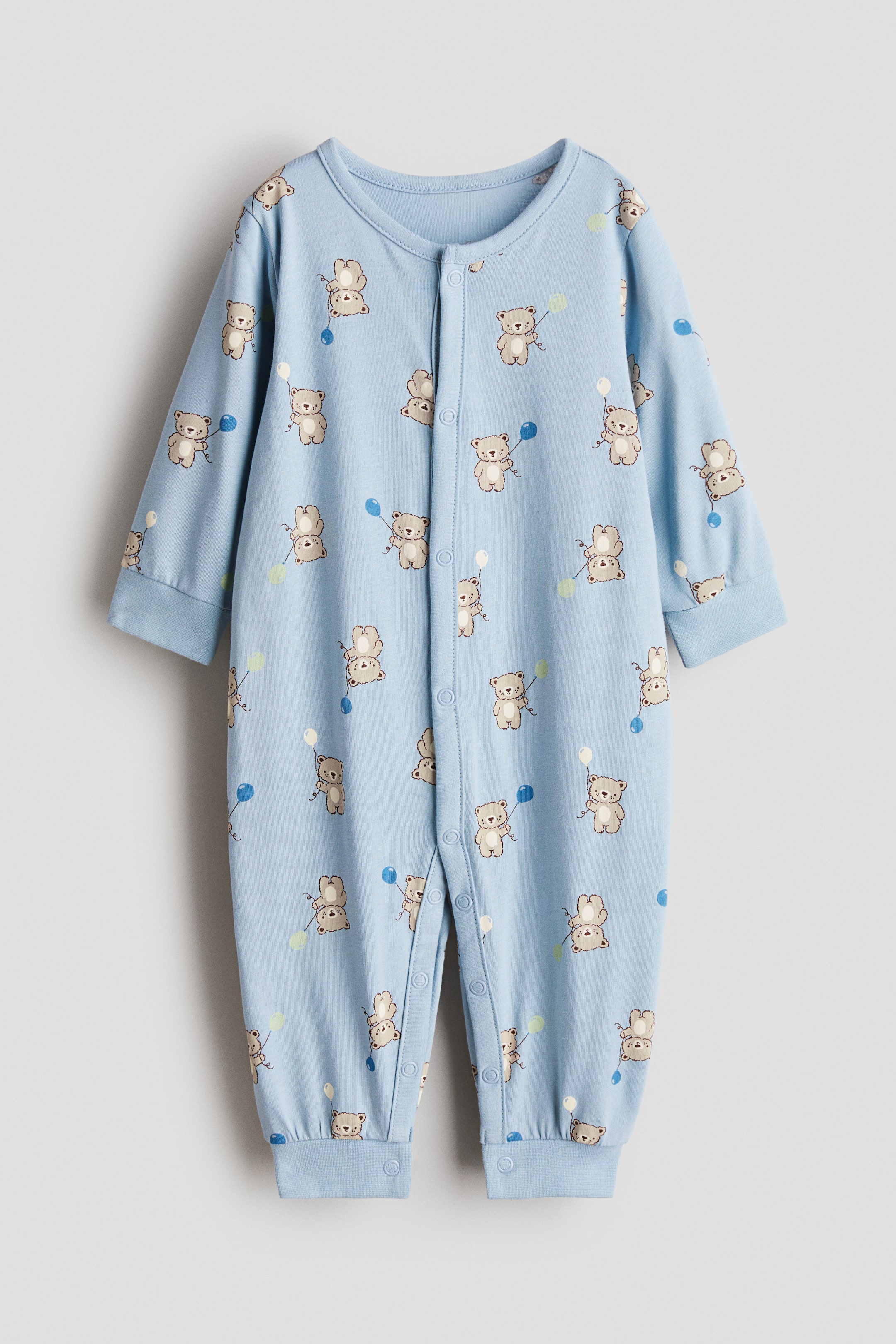 拡大画像を表示: スリープスーツのFRONTが表示されており、淡いブルーのスーパーソフトコットンジャージー製で、風船を持ったテディベアの総柄プリントが施されています。ラウンドネック、袖口と裾がカフ仕様の長袖と長めの脚部、前面と脚部に沿って並ぶプレススタッド付きでデザインされています。