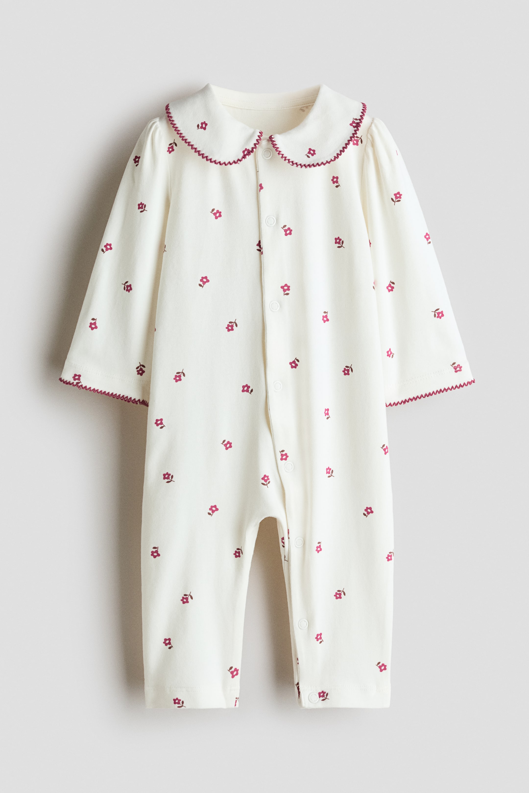 View larger image: A baby romper is presented facing front, featuring an off-white fabric with a scattered red floral print. It is designed with a Peter Pan collar and slightly puffed long sleeves, both edged with a red zig-zag stitch, and includes a full-length snap button closure extending down the front and one leg.