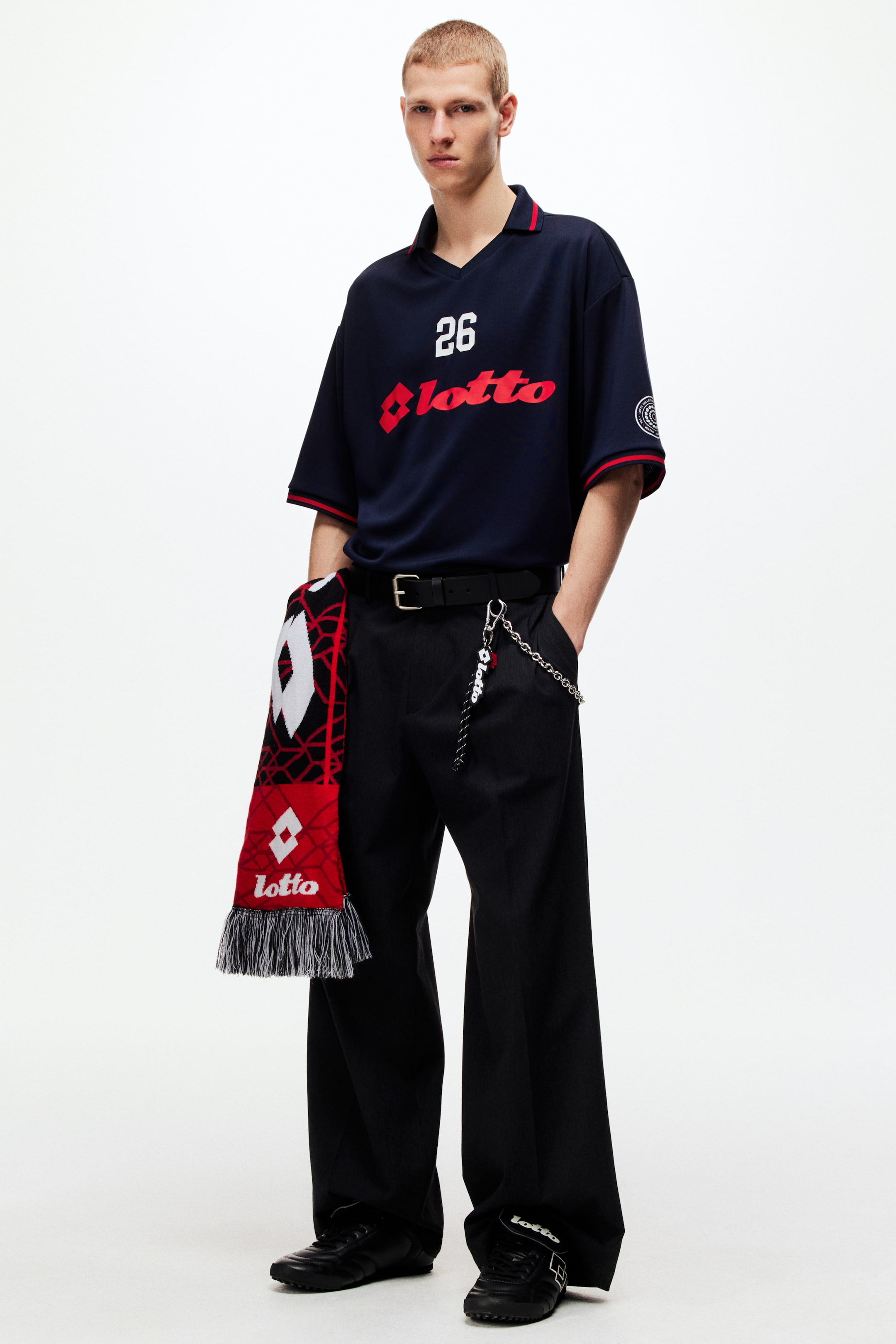 View larger image: This ensemble showcases a navy blue short-sleeved V-neck polo shirt, featuring a red and navy striped collar and cuffs. It is adorned with a white '26' and red 'lotto' logo on the chest. The look is completed with black wide-leg trousers, a black belt with a silver chain and 'lotto' keychain, black lace-up athletic shoes, and a red, black, and white knitted 'lotto' scarf with fringed ends elegantly draped over the left hip.