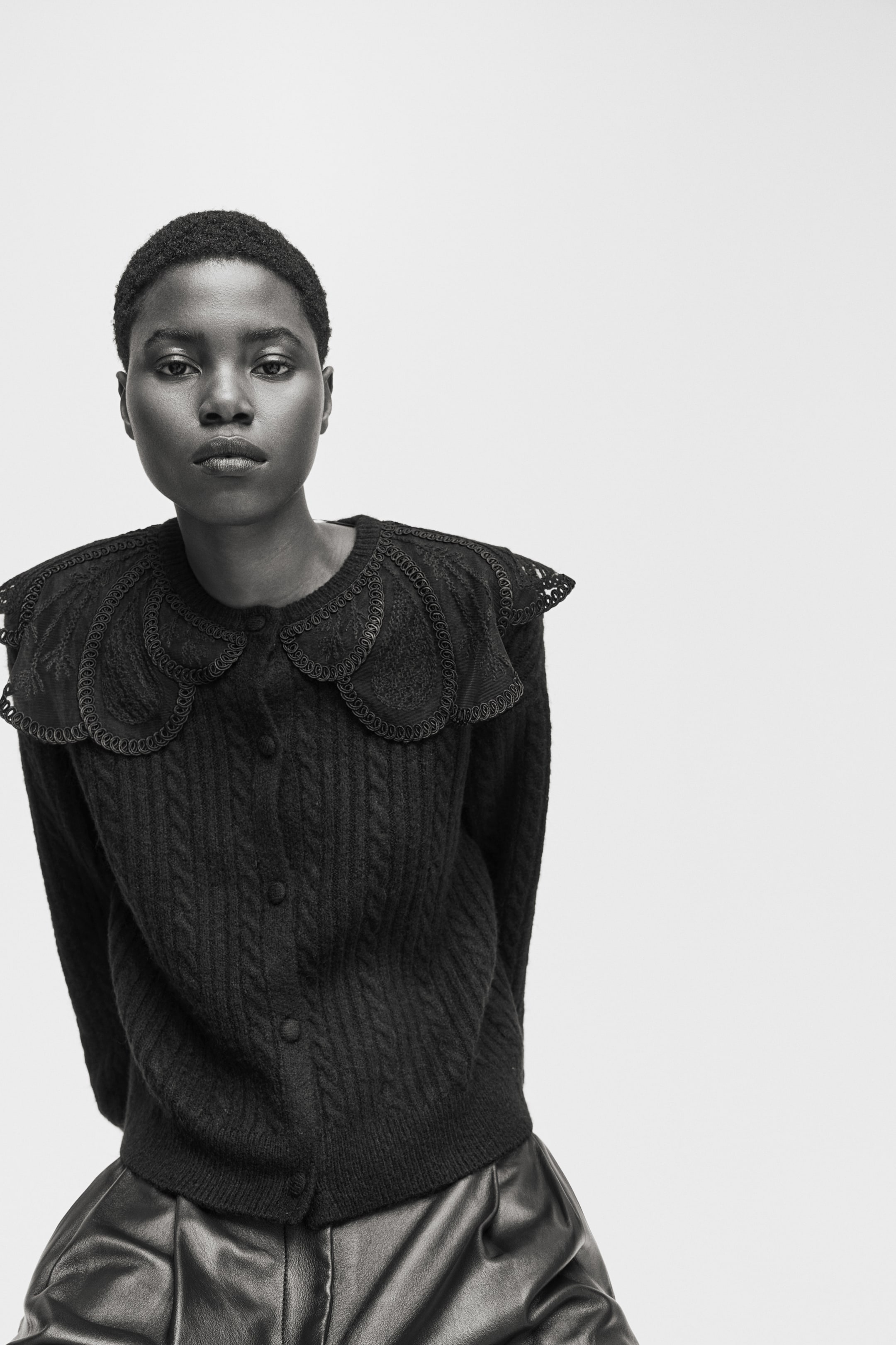 拡大画像を表示: 黒のケーブルニットカーディガンは、大きく丸みを帯びたスカラップの襟が特徴で、複雑なレースのディテールと編み込みのトリムがあり、肩にかけて広がっています。カーディガンはまた、前面にボタン留めが施されています。
