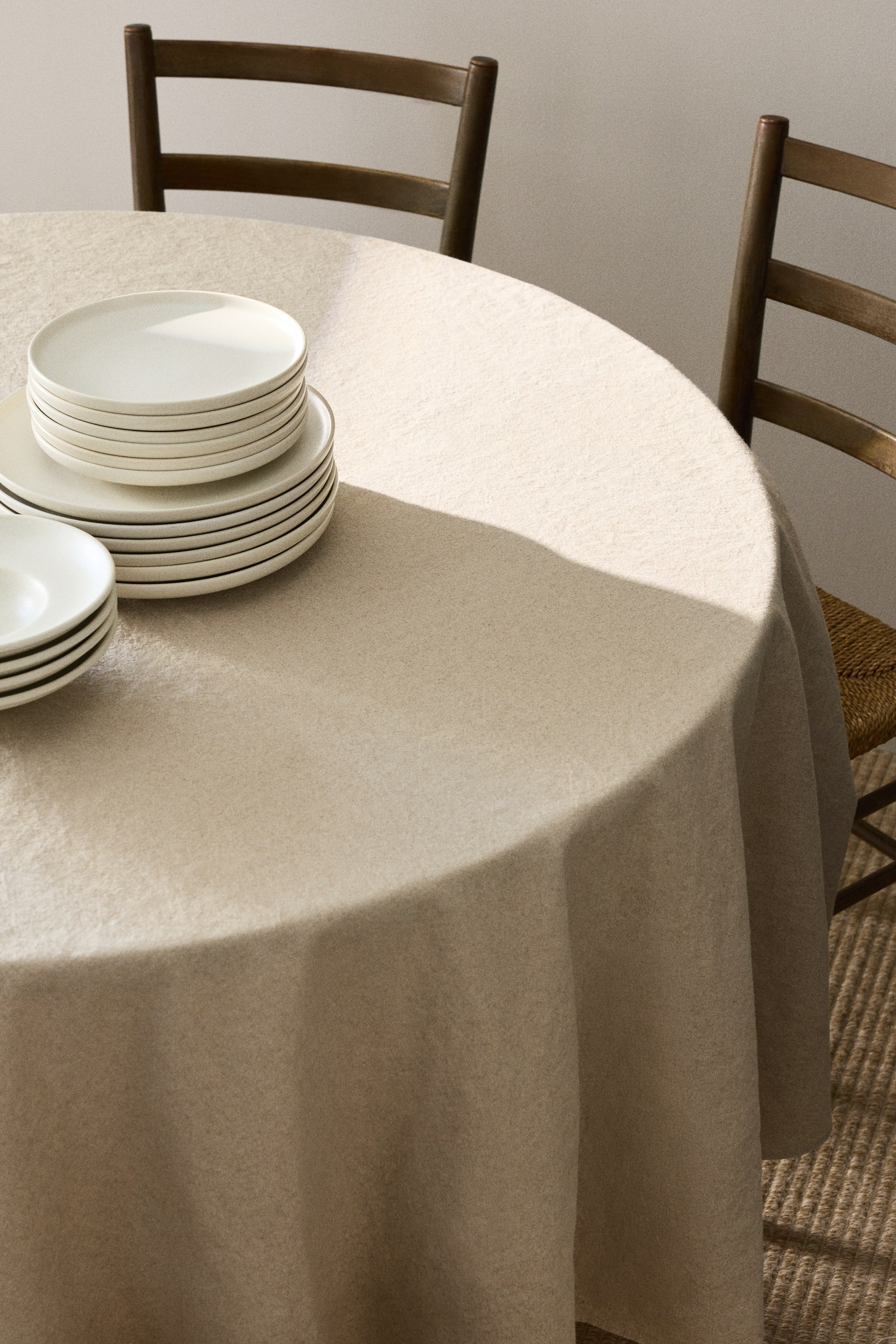 View larger image: A round tablecloth, crafted from a linen and cotton blend, drapes elegantly over a table. It showcases a light beige hue with a subtle woven texture, while stacks of white plates rest gracefully on its front surface.