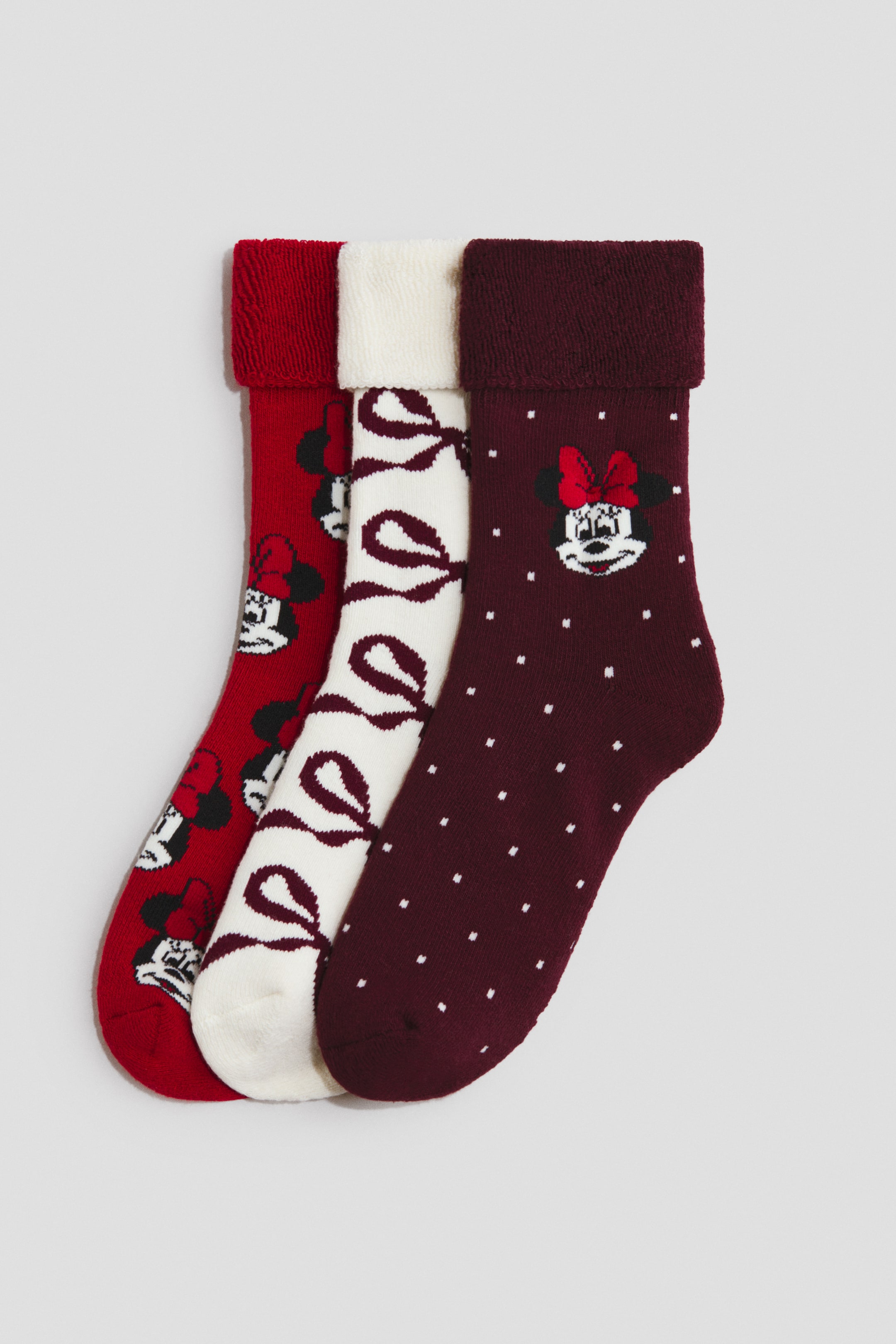View larger image: Three cozy knit socks are shown in an overlapping arrangement, each featuring a plush folded-over cuff. The first sock is red with a repeating Minnie Mouse pattern, the second is white with repeating maroon bows, and the third is maroon with white polka dots and a single Minnie Mouse graphic near the ankle.