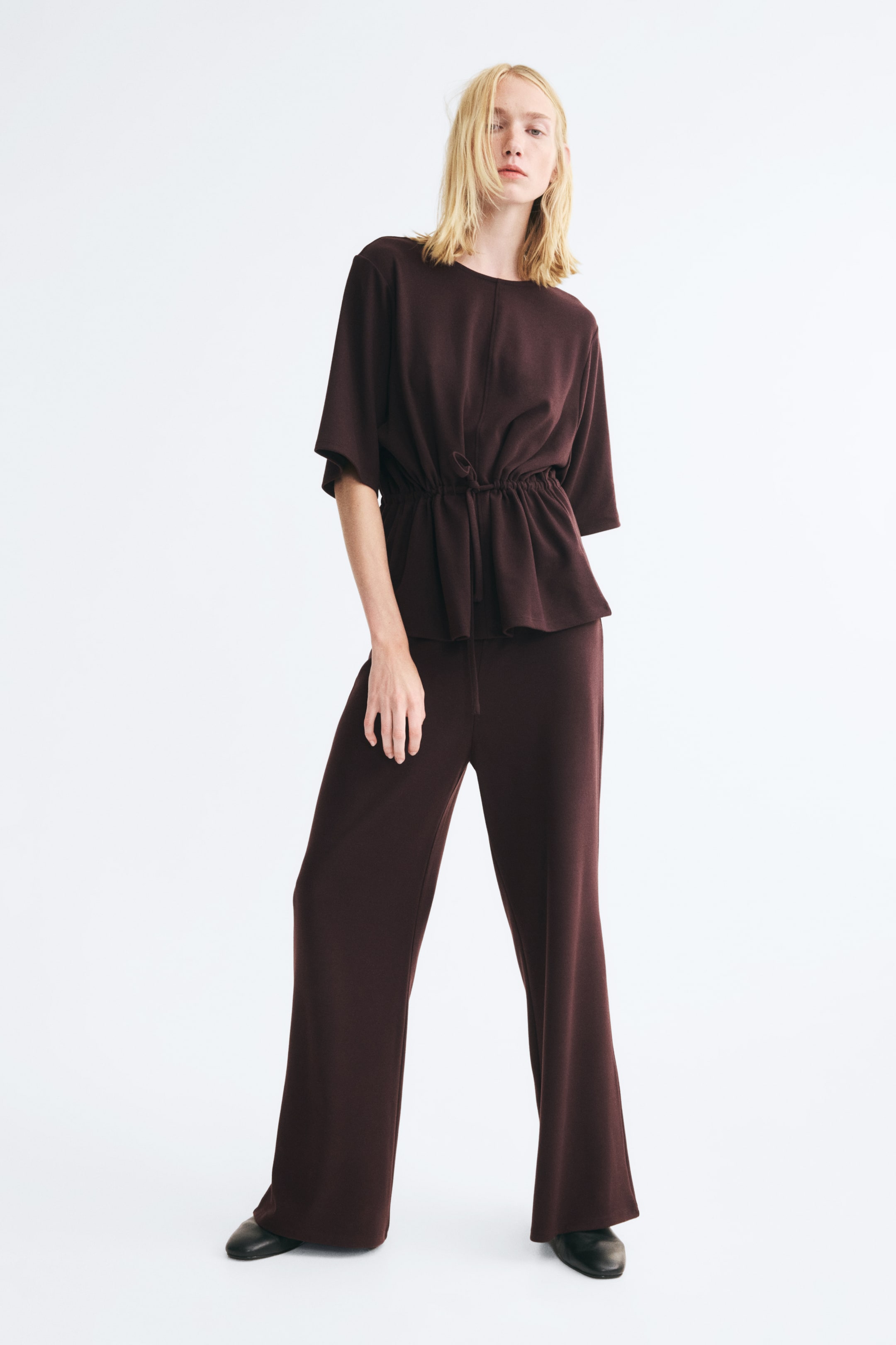 Ver imagen más grande: La modelo lleva un conjunto a juego de punto en marrón oscuro, compuesto por un top de manga corta con cuello redondo y cintura fruncida con cordón ajustable, combinado con unos pantalones de pernera ancha a juego y zapatos negros.