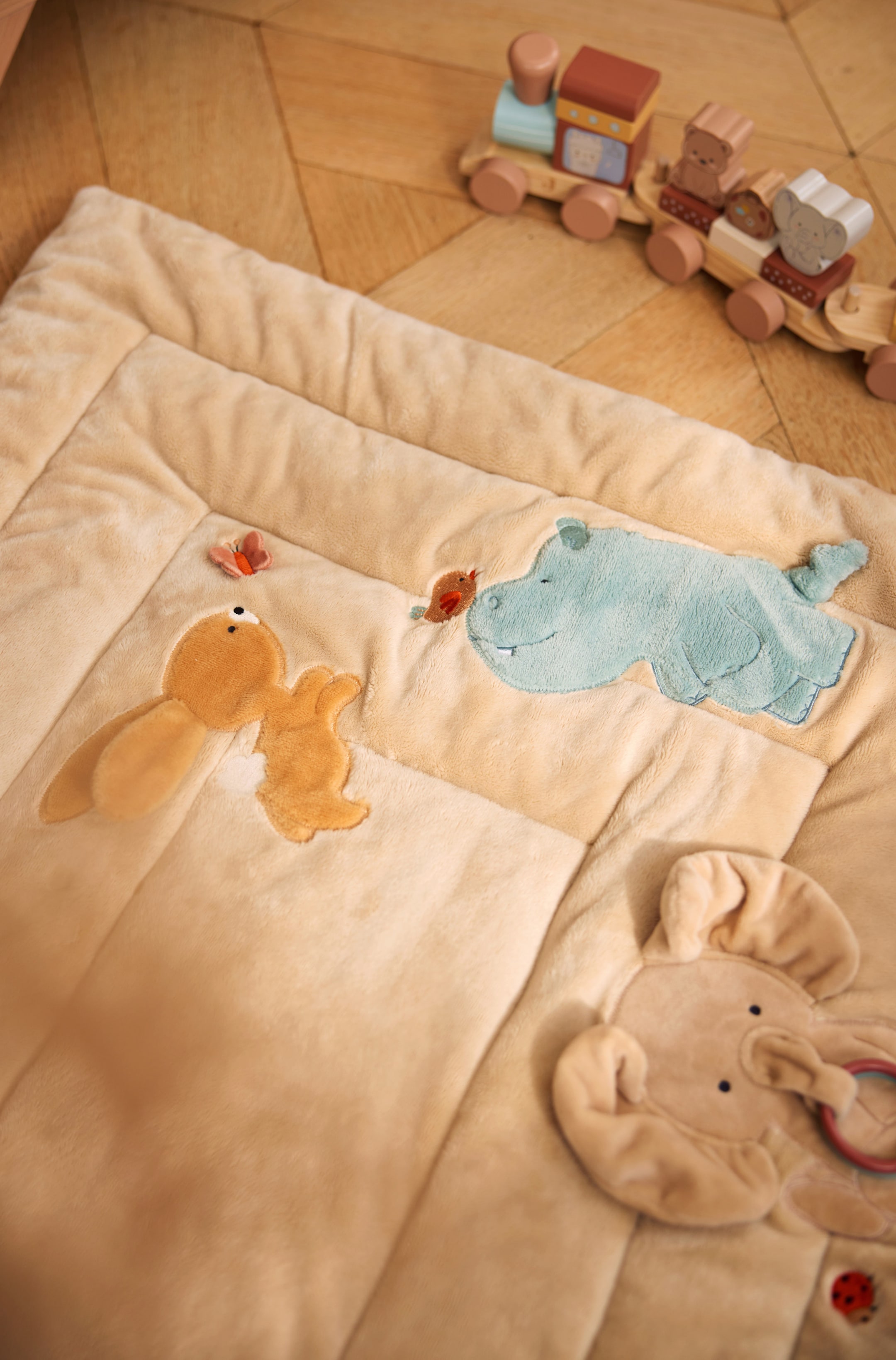 Größeres Bild ansehen: Eine weiche, gepolsterte beige Baby-Spielmatte mit gesteppter Optik und sanft vernähten Rändern. Sie zeigt verschiedene erhabene Tierapplikationen, darunter ein hellblaues Nilpferd, ein braunes Kaninchen und einen plüschigen Elefantenkopf, der einen roten Beißring hält.