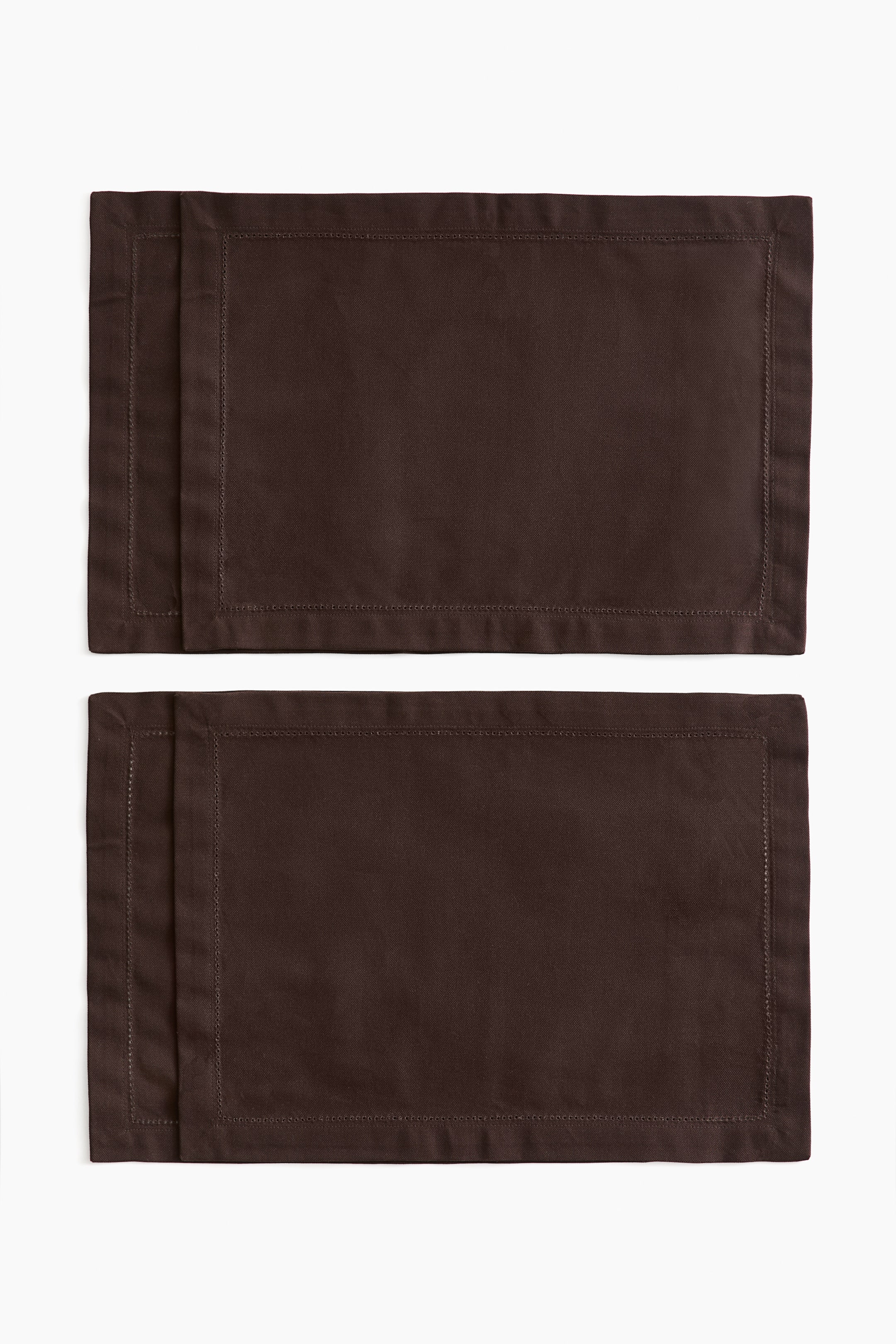 Grösseres Bild ansehen: Dunkelbraune Tischsets aus Baumwoll-Canvas liegen flach in zwei leicht versetzt übereinanderliegenden Paaren. Jedes rechteckige Tischset zeigt einen breiten Saum mit feinen Saumstich-Details entlang der inneren Bordüre.