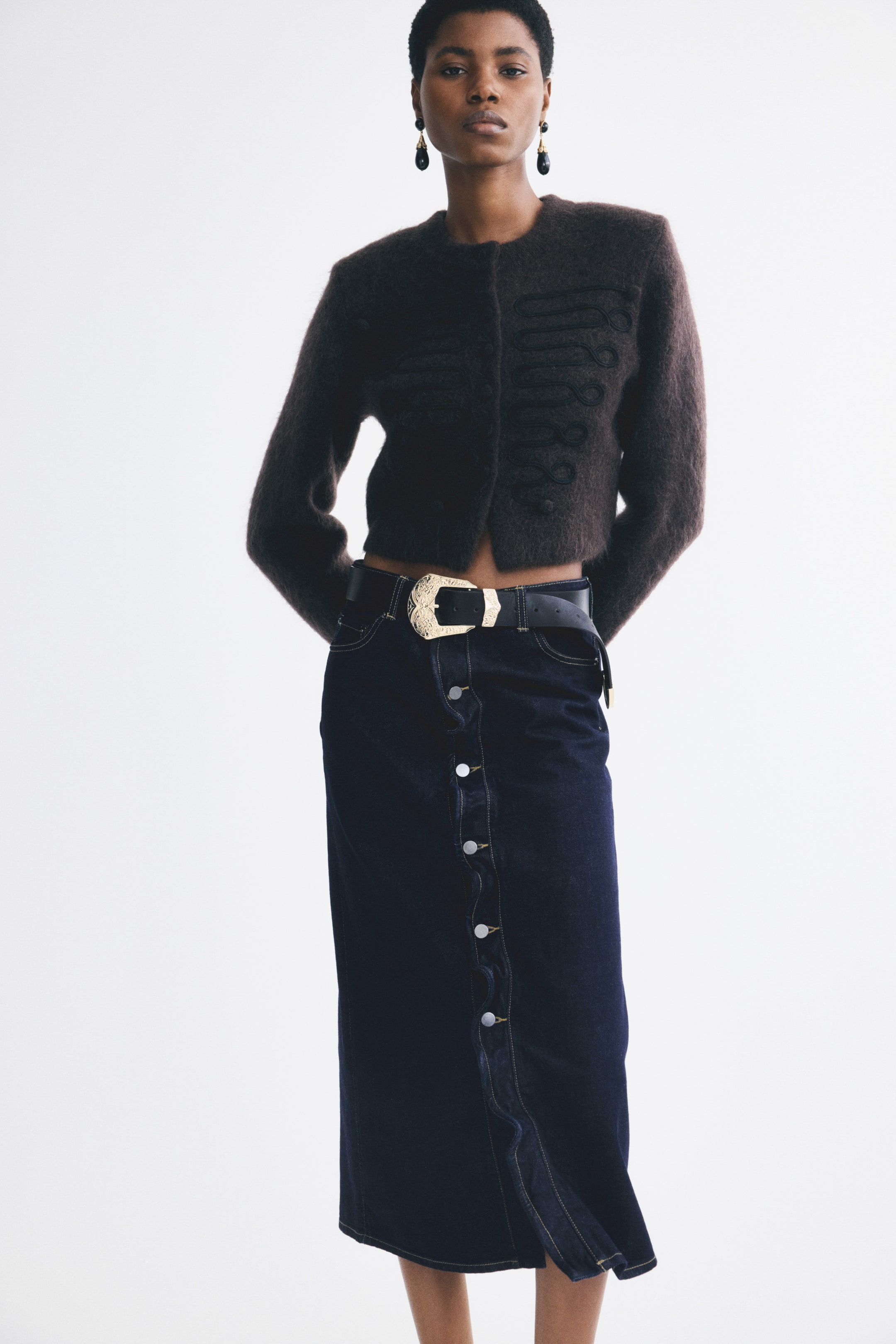 View larger image: A calf-length, dark blue denim skirt featuring a button-down front with ruffled trim is being worn by the model. The skirt is styled with a wide black belt featuring an ornate gold buckle and paired with a cropped, fuzzy brown cardigan.