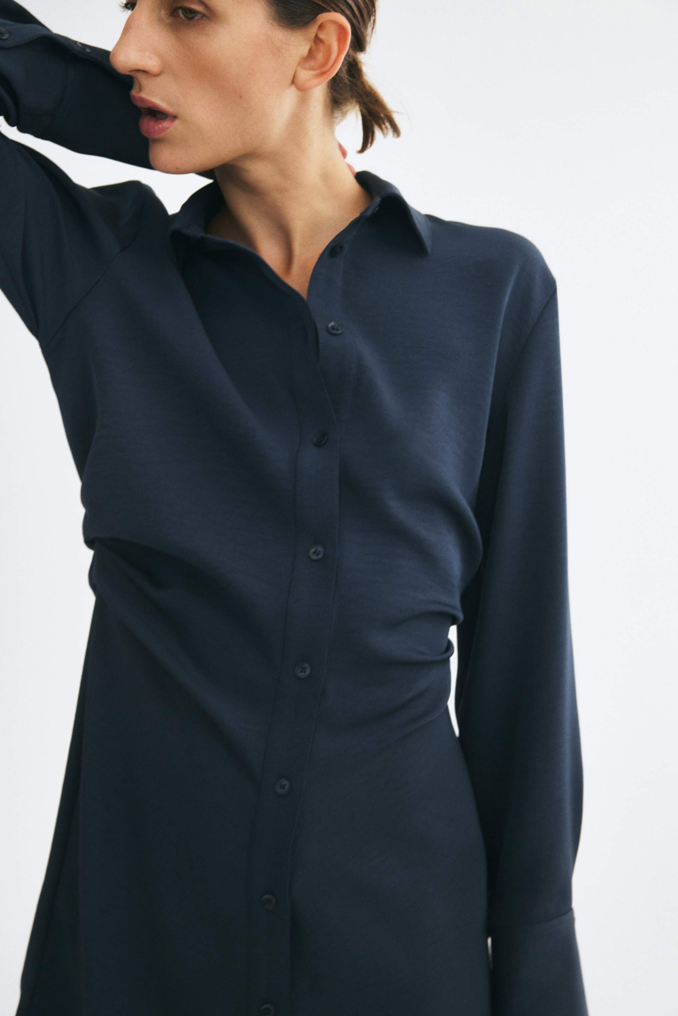 Ampliar la imagen: La modelo lleva un vestido camisero ajustado de color azul oscuro, con cuello, cierre de botones al frente y detalle fruncido en la cintura. Posan con un brazo levantado hacia la parte posterior del cuello, resaltando las mangas largas y la silueta drapeada.