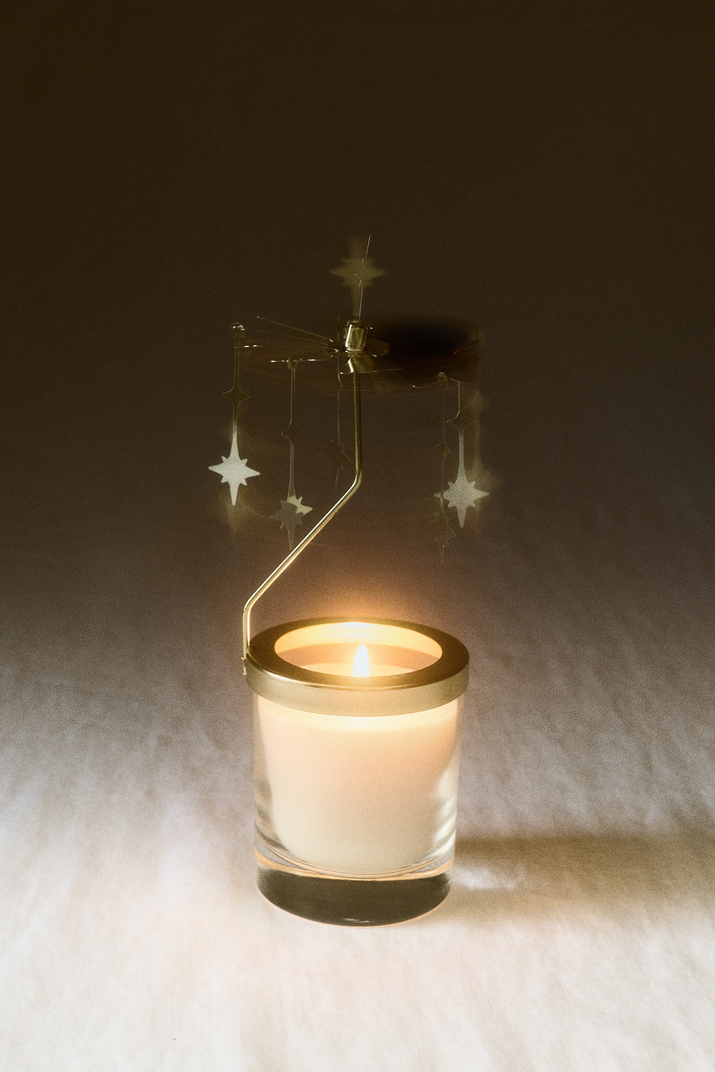 Un set de carrusel para vela, mostrado de frente, presenta una vela blanca en un frasco de vidrio transparente junto con su caja de empaque rectangular de color claro. El accesorio de carrusel color dorado descansa sobre el borde del frasco con dijes de estrella colgantes, mientras que la caja a juego presenta lÃ­neas en foil dorado y patrones de estrellas.