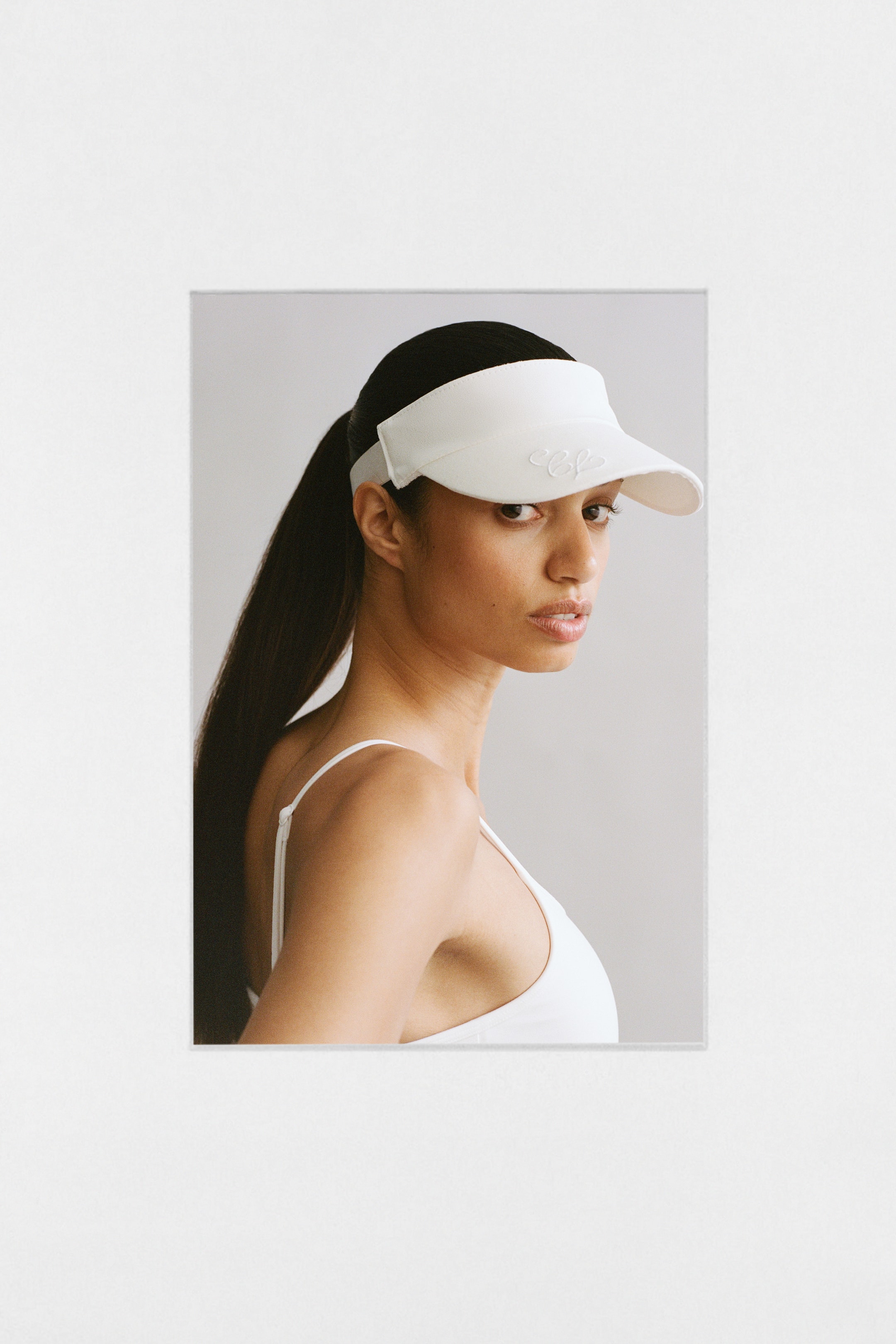 Agrandir l'image: Le modèle porte une visière blanche avec une visière incurvée et un logo brodé, ses cheveux foncés étant tirés en arrière à travers l'ouverture.