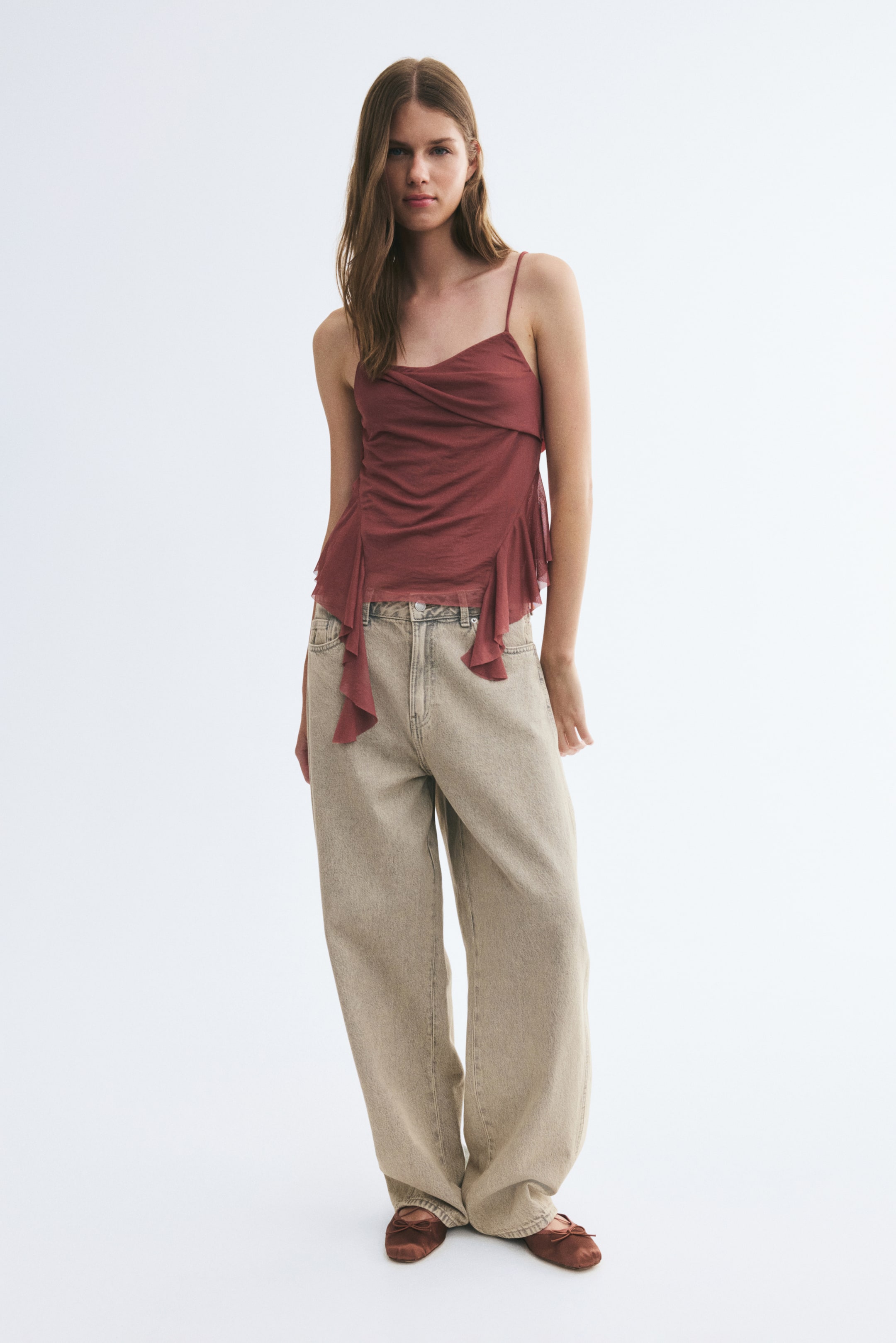 View larger image: A sheer mesh cami in dark rose is worn by the model, featuring thin spaghetti straps, an asymmetrical foldover front, and cascading flounced ruffles along the sides of an uneven hem. The top is styled with light beige wide-leg jeans.