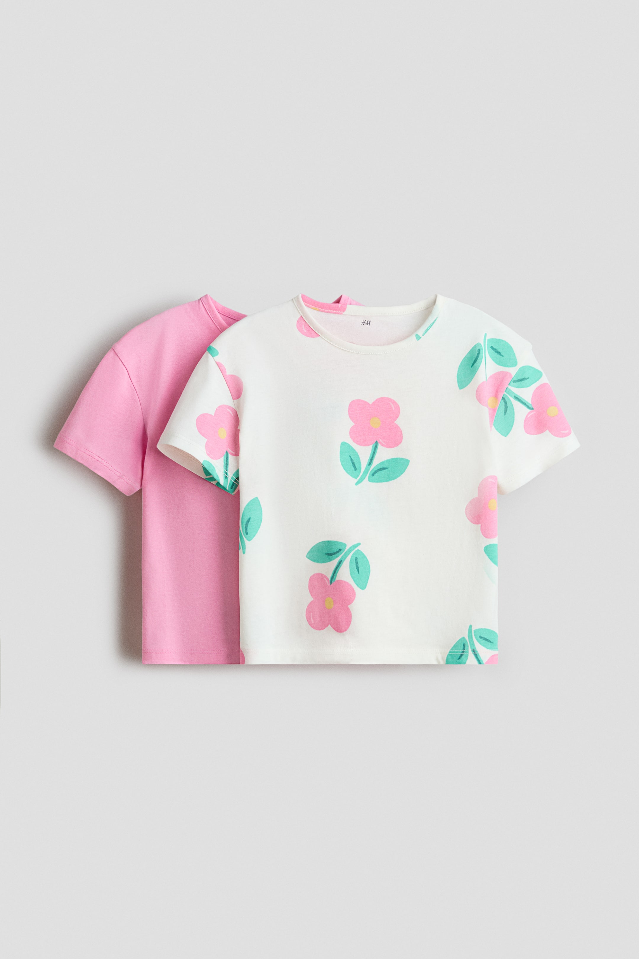 Größeres Bild ansehen: Das vordere T-Shirt ist cremeweiß und zeigt ein sich wiederholendes Muster aus rosa Blumen mit gelben Zentren und türkisfarbenen Blättern. Es ist mit einem Rundhalsausschnitt und kurzen Ärmeln gestaltet, und ein einfarbiges rosa T-Shirt mit Rundhalsausschnitt und kurzen Ärmeln ist teilweise dahinter sichtbar.