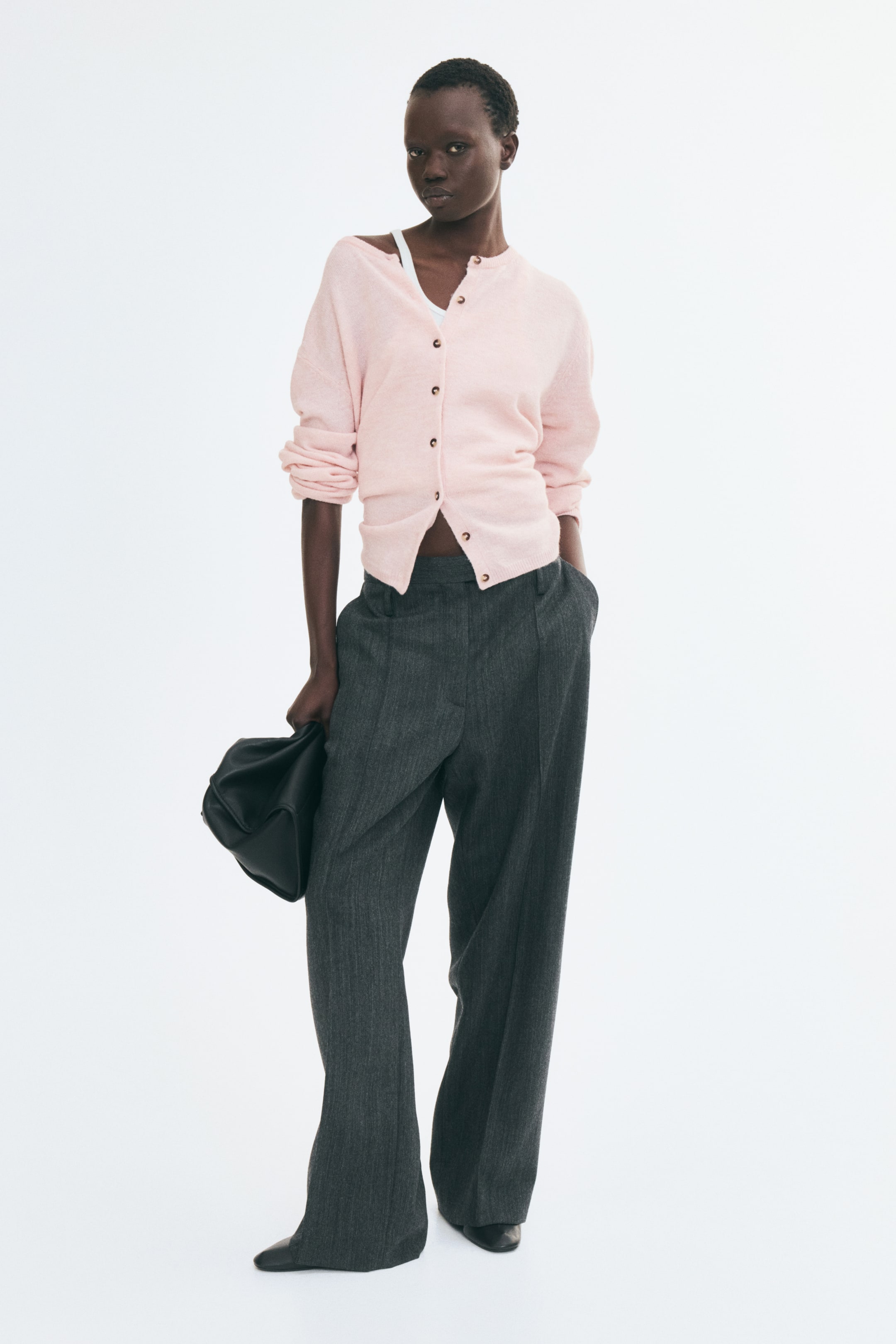 View larger image: A light pink, soft-knit cardigan is being worn by the model, featuring a partially buttoned front and a round neckline draped slightly off one shoulder. The cardigan is being layered over a white top and styled with dark grey wide-leg trousers, with its long sleeves pushed up on the arms.