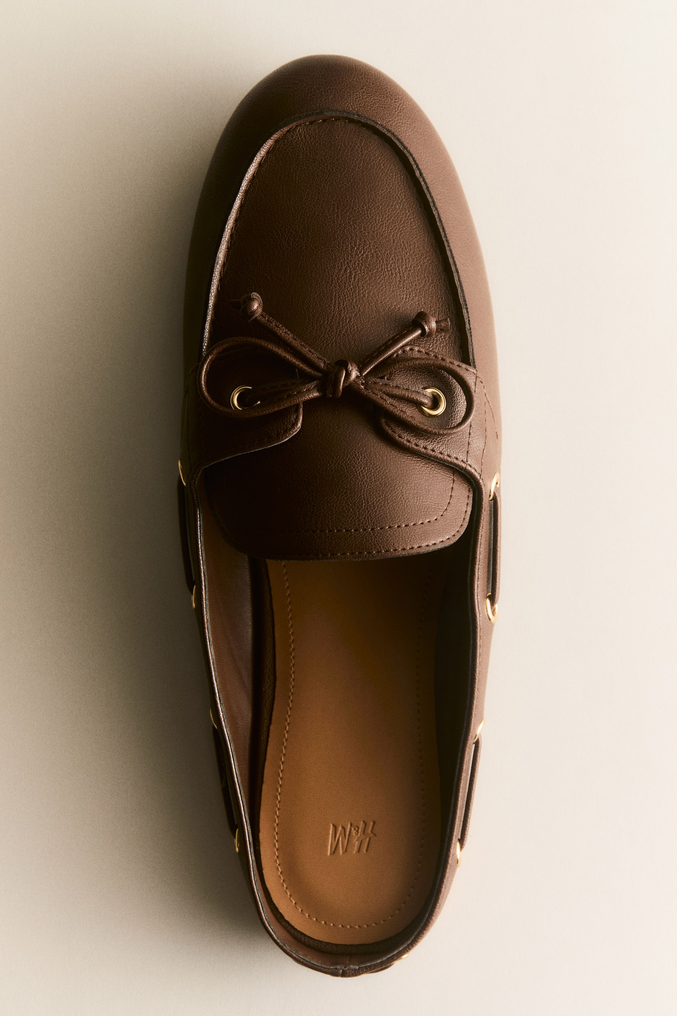 View larger image: Presenting a dark brown leather loafer mule, showcasing a moccasin-stitched vamp complemented by a decorative bow threaded through two gold-toned eyelets. Additional gold-toned eyelets embellish the sides, enhancing its sophisticated appeal. The light brown insole, embossed with "H&M," is distinctly visible within the shoe, adding a touch of elegance.