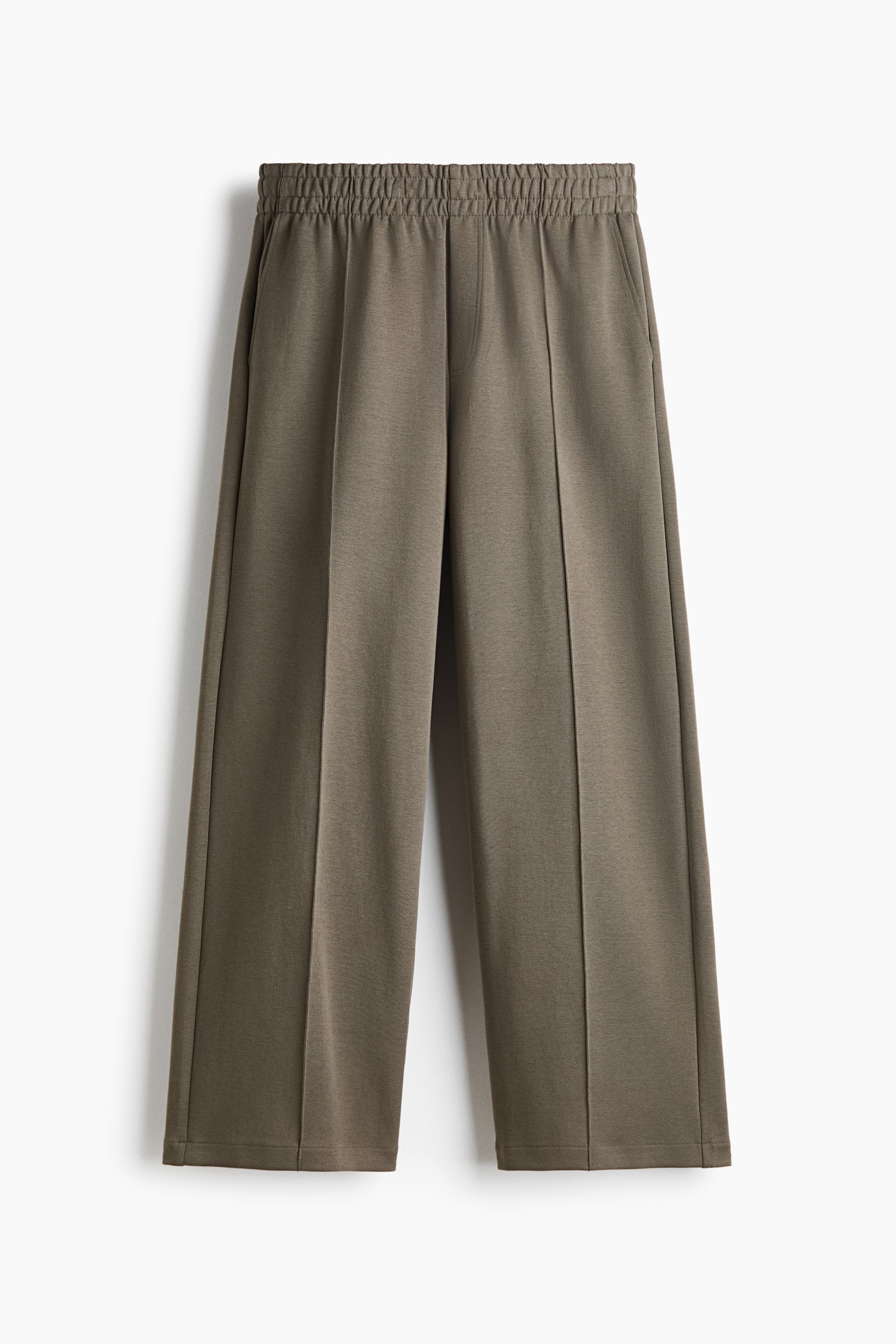A pair of taupe-coloured wide-leg trousers is shown from the front, featuring a gathered elastic waistband and a stitched mock fly. The trousers display permanent stitched creases running down the centre of each leg.