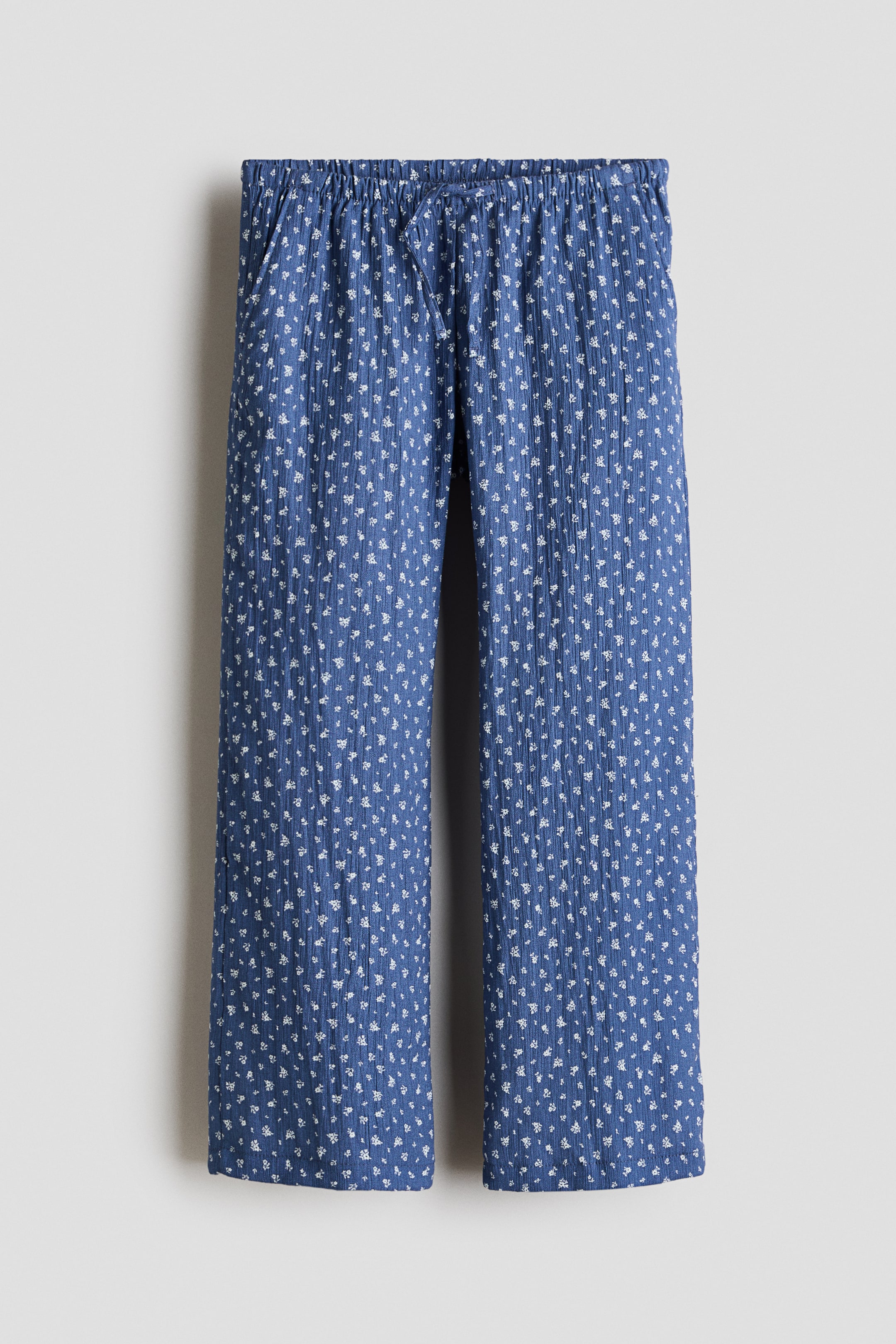 View larger image: Relaxed-fit trousers in blue woven fabric with a white floral pattern are displayed from the front, featuring an elasticated, drawstring waist with visible ties and diagonal side pockets.