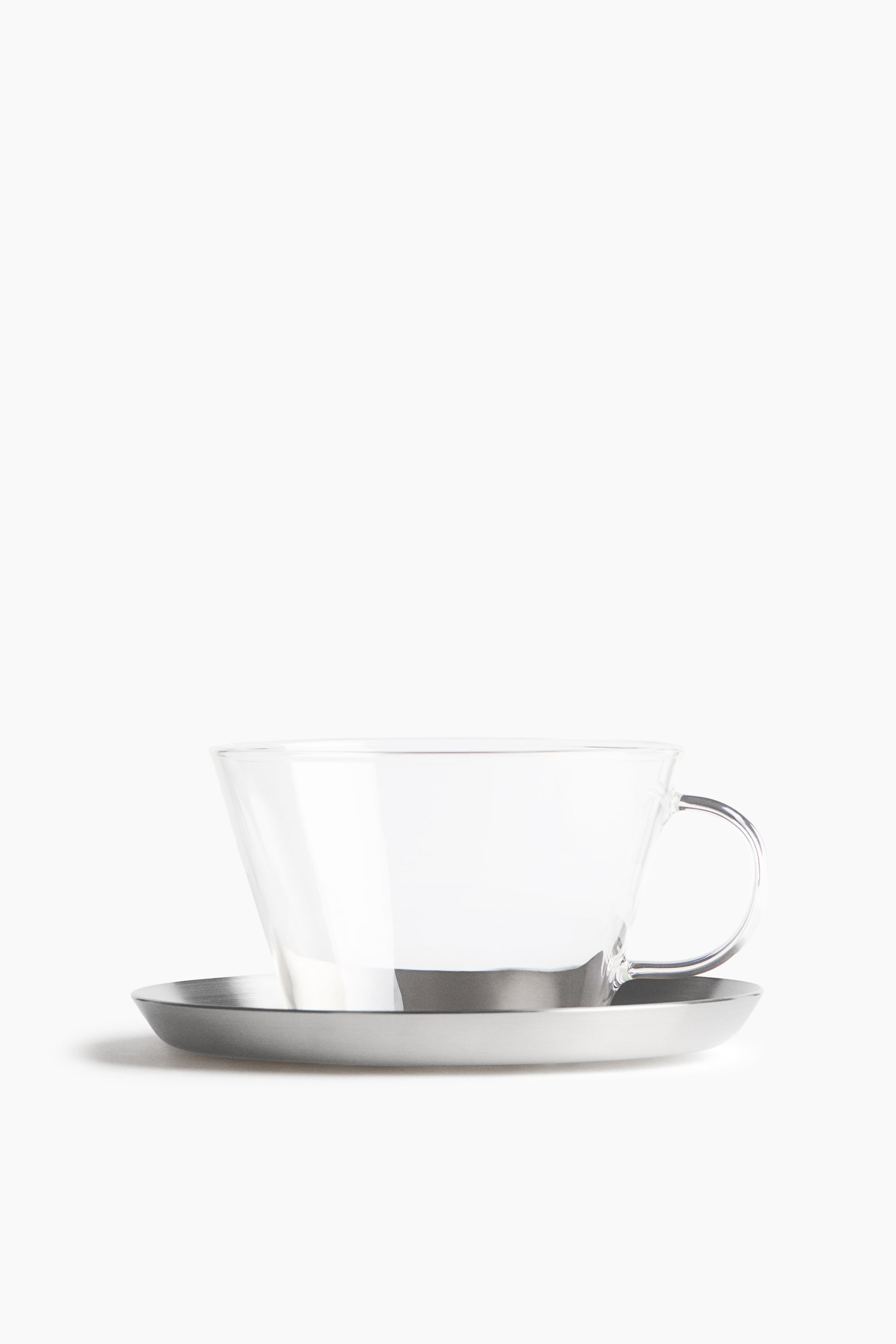 View larger image: A clear glass teacup with a flared rim and a curved handle is resting on a shallow brushed stainless steel saucer, facing forward.