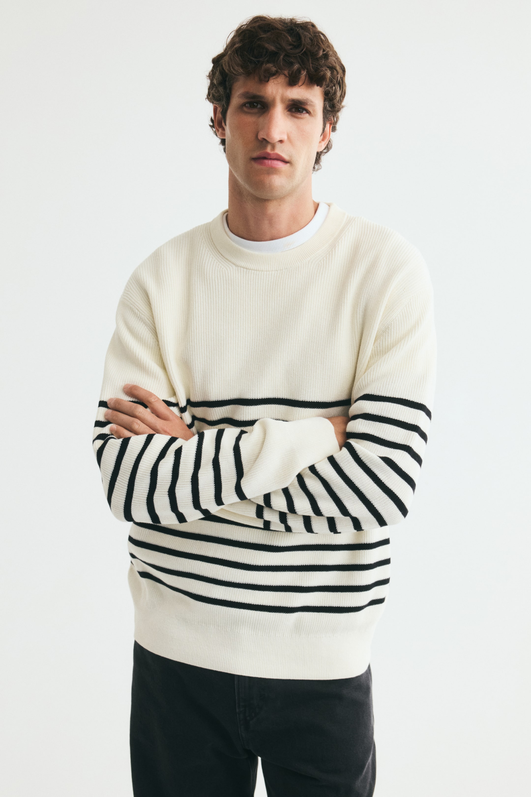 Se større billede: En ribstrikket bomuldstrøje i råhvid med vandrette sorte striber bæres af modellen, som står med armene over kors. Trøjen i regular fit har rund halsudskæring, der viser en hvid T-shirt i lag under, sænkede skuldre og lange ærmer.
