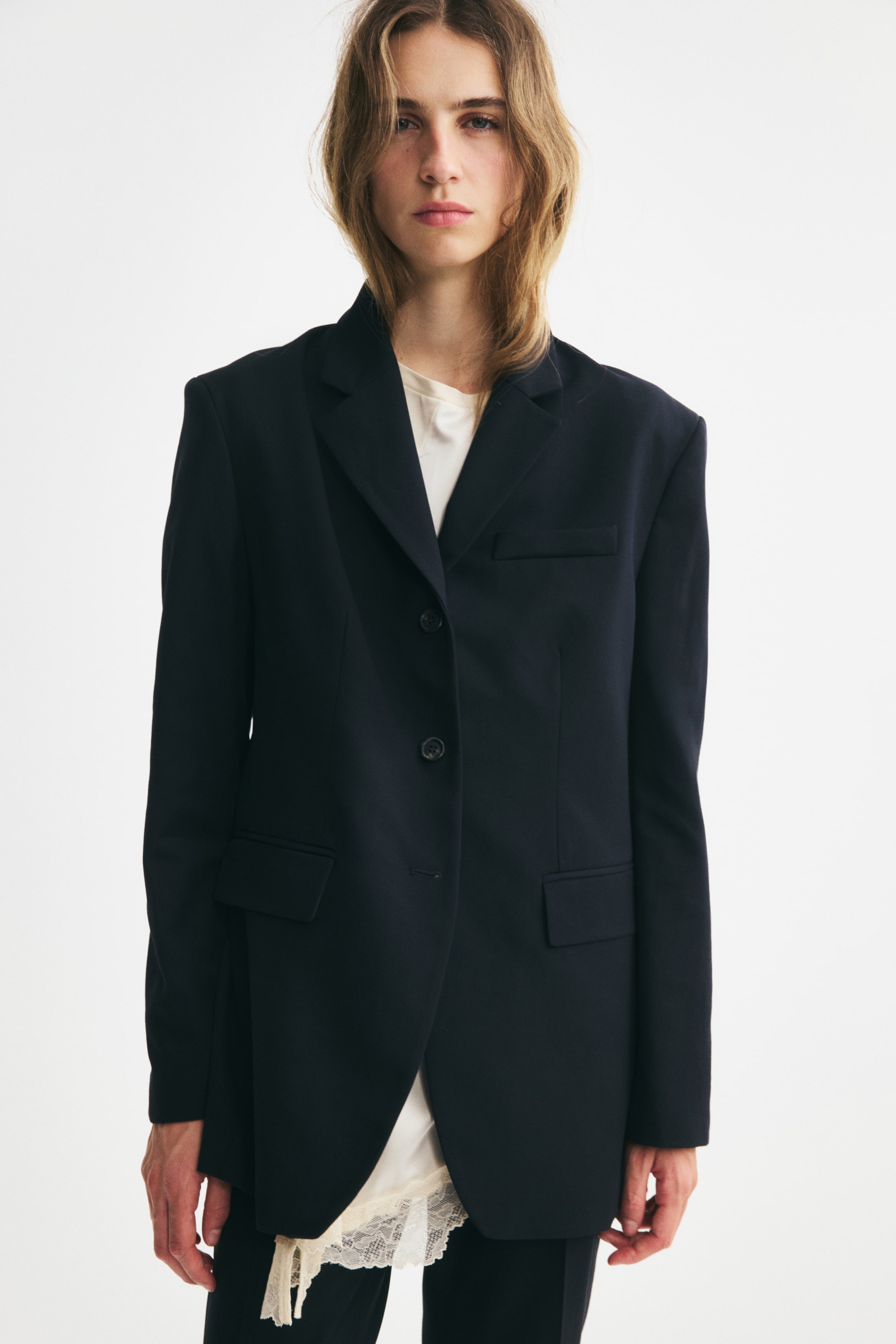 View larger image: A long, single-breasted black twill blazer is shown from the front, featuring structured shoulders, notch lapels, and waist darts that create a tailored silhouette. The blazer is designed with a welt chest pocket, two front flap pockets, and a three-button closure, styled over a cream top with a visible lace hem.