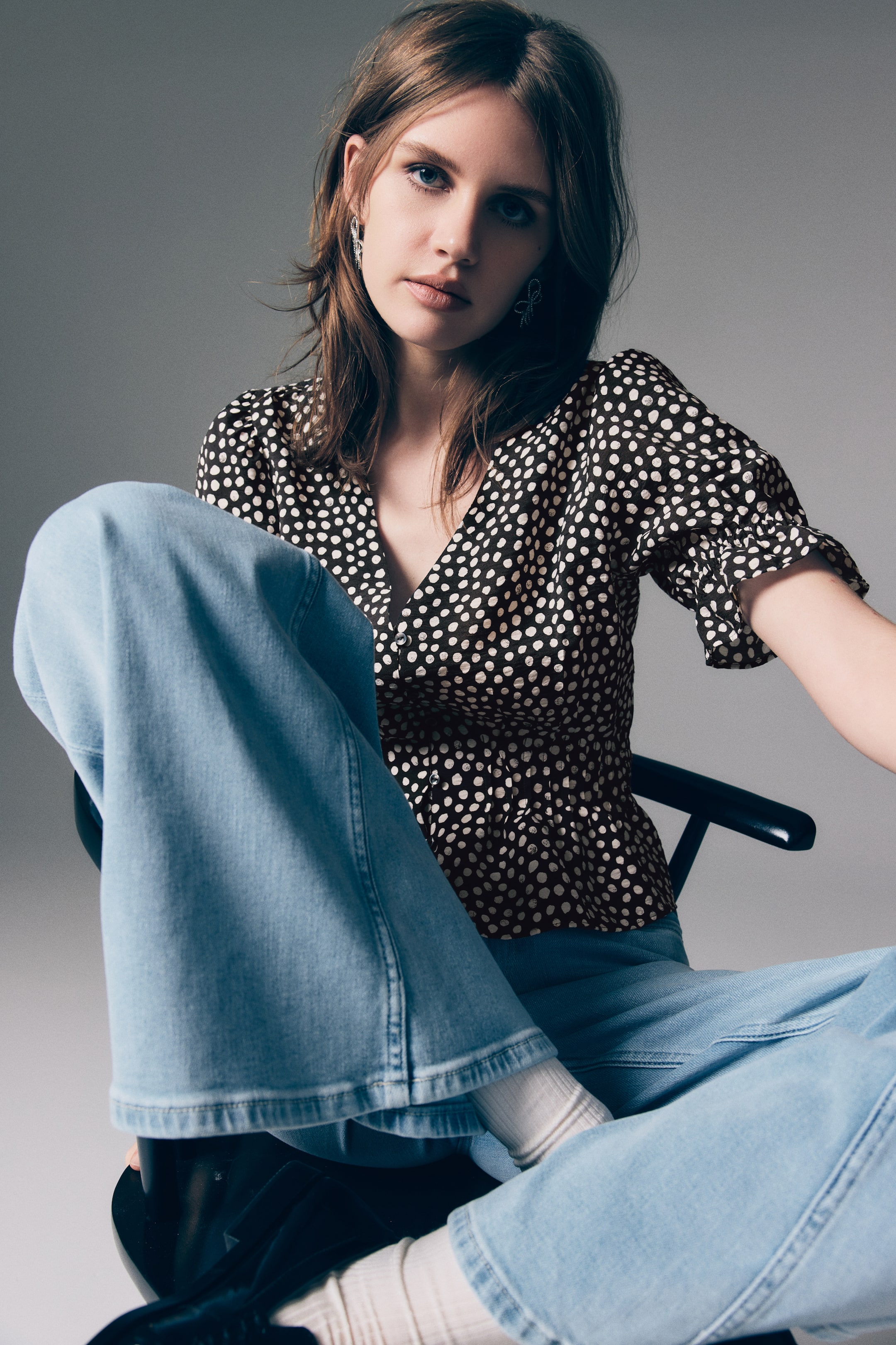 View larger image: A black top with irregular white polka dots is featuring a V-neckline and a wrap-style front with visible buttons. It is presenting short, gathered sleeves with ruffled cuffs and a cinched waist leading to a peplum hem.